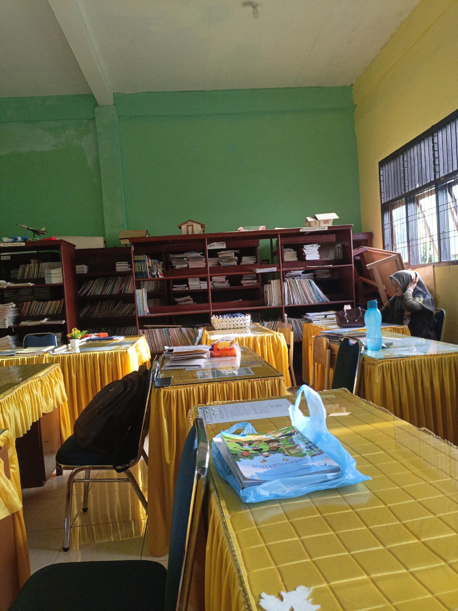 The image depicts the interior of a classroom or library in Lhokseumawe, Indonesia. The room features bright green and yellow walls. Several tables are covered with yellow tablecloths. On one table, a stack of books is placed inside a blue plastic bag. A backpack is on a chair. Bookshelves line the walls, filled with numerous books. A person wearing a headscarf is seated at a table in the background, near a window with bars.