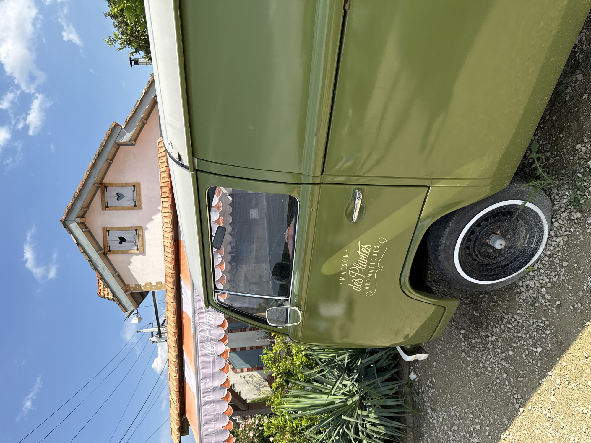 A vintage green van is parked on a gravel and dirt surface in front of a building with a striped awning. The van has a logo on its side that reads "MAISON des PLANTES AROMATIQUES" in white lettering. The building behind the van appears to be a shop or restaurant, with a light pink facade and a tiled roof. There are two windows on the upper story of the building, each with a shutter cut into a heart shape. Lush green foliage and a tall, spiky plant are visible to the left of the van. The sky above is a clear blue with a few white clouds. The scene is bathed in sunlight, casting shadows on the ground. The location is identified as Semogorskiy, Russia.