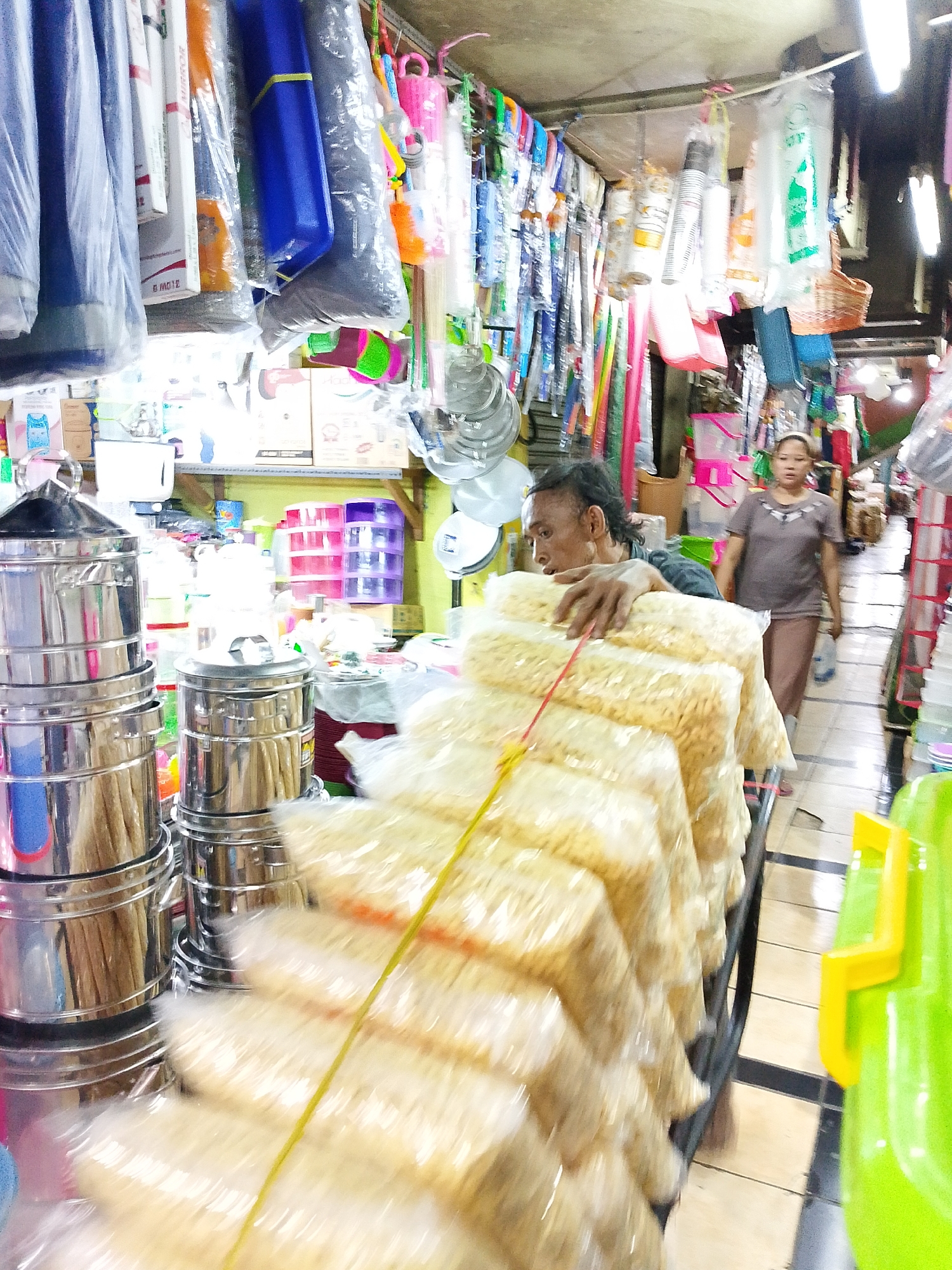 This is a blurry, medium-shot photograph taken indoors, likely in a market stall in Surabaya, Indonesia. The scene is crowded with merchandise, primarily household goods and packaged food items. In the foreground, a man with dark, slicked-back hair and a concerned expression is leaning over a display of large, clear plastic bags filled with light-colored, puffed snacks. He is wearing a dark blue-grey shirt and appears to be arranging or presenting the goods. A yellow string runs across the bags.
Behind the man, various stacked metal containers, possibly steamers or pots, are visible. To the right of the man, a woman in a taupe-colored shirt and pants is walking away from the camera, carrying a white plastic bag in her right hand. She has dark hair and a neutral expression. The background is filled with hanging items, including colorful plastic buckets, cleaning brushes, and more packaged goods, suggestive of a busy retail environment. Shelves laden with boxes and containers are also visible. The lighting is somewhat harsh, and the overall image has a motion blur, indicating movement during the capture. The floor appears to be tiled.