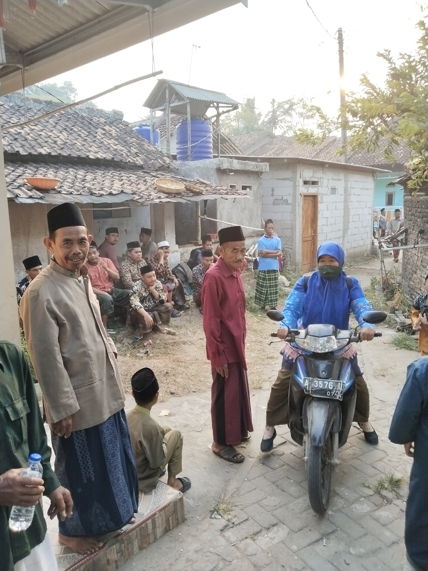 In Serang, Indonesia, on a dusty, unpaved street lined with modest, tile-roofed houses, a group of men are gathered. The foreground is dominated by a man in a beige jacket and patterned sarong, who smiles at the camera. To his left, another man, partially visible, holds a bottle of water. A young boy in a tan tunic and black peci (cap) sits on a step to the right. Further back, a woman wearing a blue hijab and face mask rides a motorcycle. Other men, dressed in traditional attire including pecis and sarongs, are seen sitting and standing in the background. The scene appears to be a casual gathering, possibly a community event or a break during a day's activities, with a backdrop of traditional Indonesian village life. The lighting suggests it is late afternoon, with the sun casting long shadows.