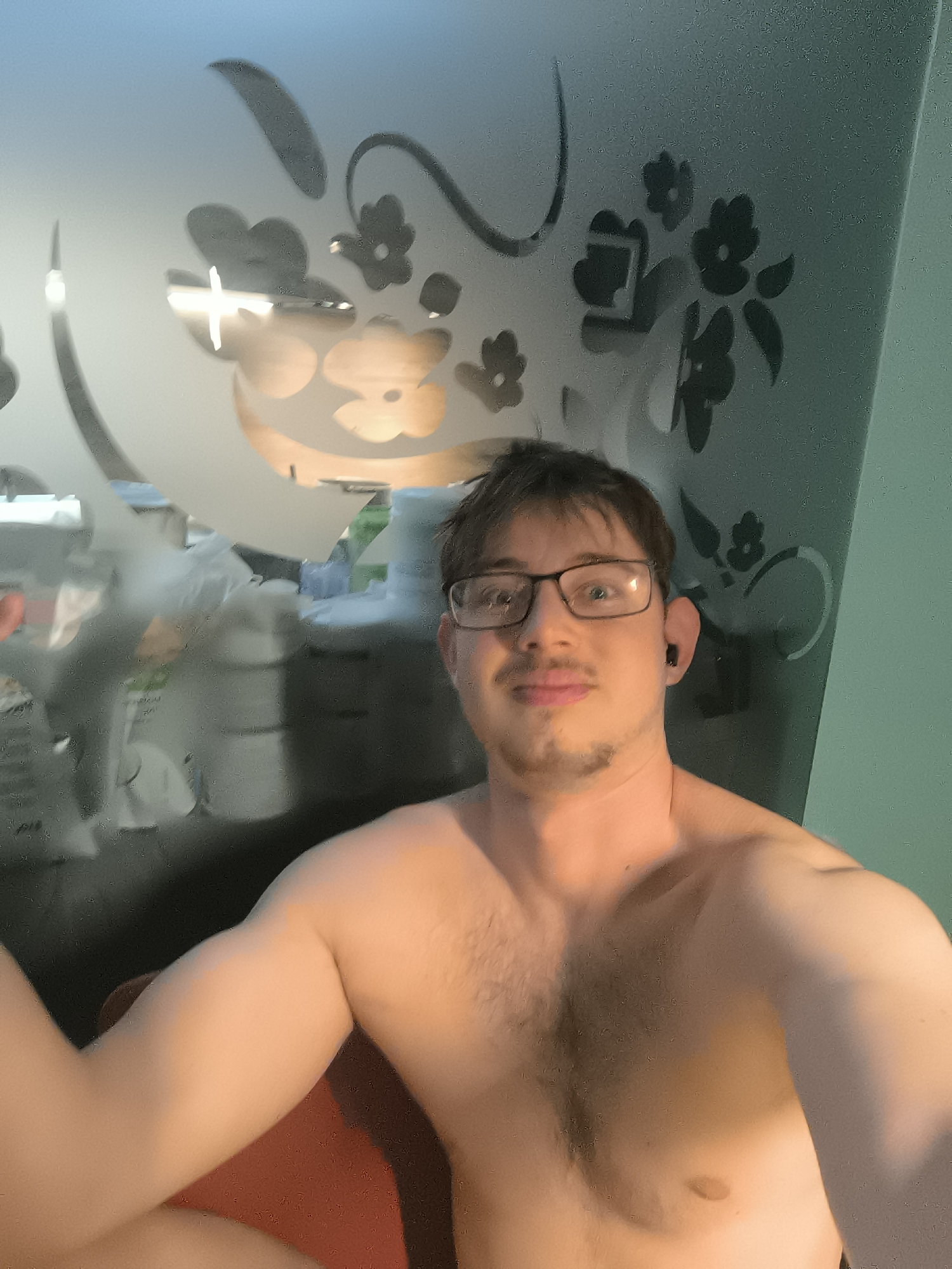 A young man with short brown hair, wearing black-rimmed glasses, and a dark earbud in his right ear, is shown in a selfie. He has light facial hair (stubble/goatee) and is shirtless, revealing a hairy chest. He looks directly at the camera with a neutral to slightly smiling expression.
The man is positioned indoors, with a prominent translucent screen or partition behind him. This screen is decorated with opaque floral and swirling patterns in a darker tone against its lighter, semi-transparent surface. Through the blurry areas of the screen, indistinct objects are visible in the background, suggesting another room or area beyond. These blurred shapes could be furniture or other household items. The lighting appears to be indoor or artificial. A hint of an orange surface, possibly a chair or cushion, is visible in the lower foreground. There are no specific visual cues within the image to confirm its location in Sofia, Bulgaria, nor is any text visible.