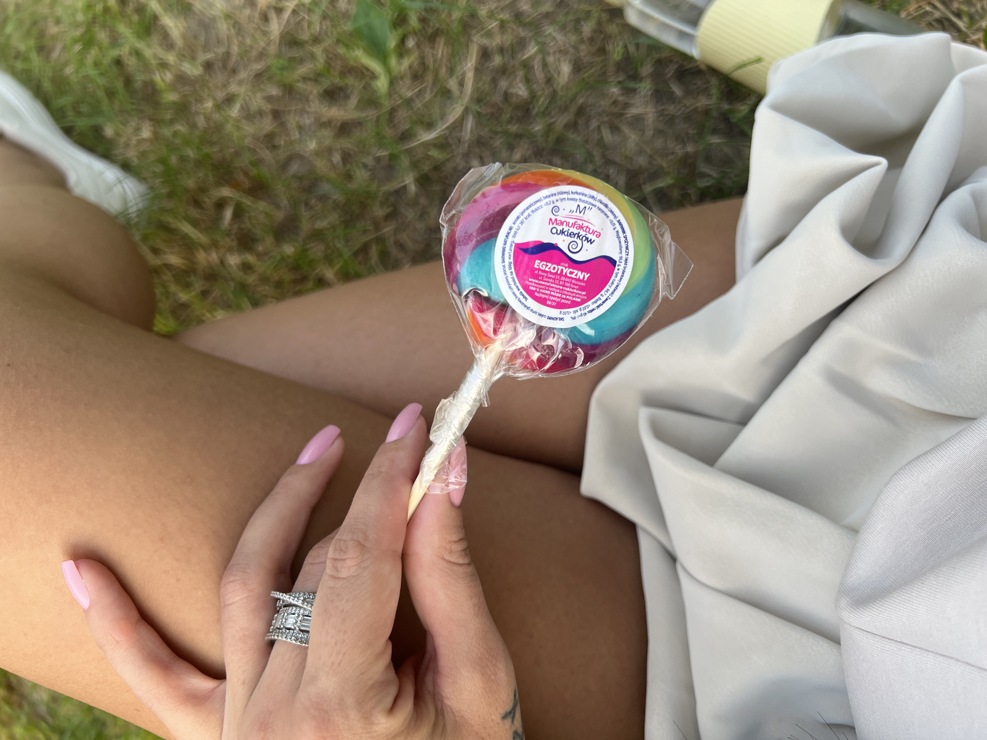 The image shows a close-up of a person's hand holding a rainbow-colored lollipop. The hand has light pink painted fingernails and is wearing a silver ring with square-cut stones on the ring finger. A tattoo is partially visible on the wrist. The lollipop is wrapped in clear plastic with a pink and blue label that includes Polish text. The person is wearing a light-colored skirt or dress. The background consists of green grass, suggesting an outdoor setting. The provided information indicates the image was taken in the city of Tychy, Poland, but there are no specific landmarks or details within the image to confirm this location.