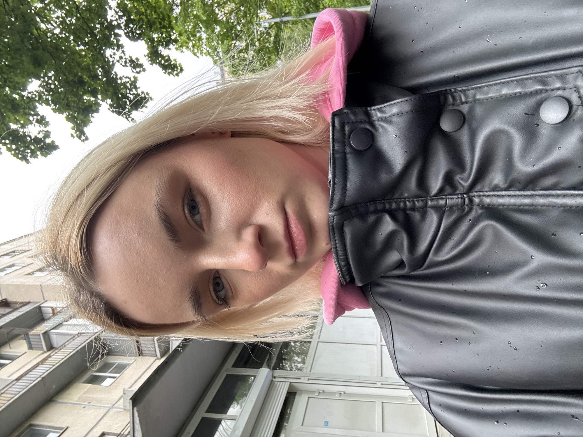 The image is a selfie of a young woman with blond hair, blue eyes, and fair skin. She is wearing a pink hoodie underneath a black leather jacket with water droplets on it. The photograph appears to have been taken outside. The background includes a building with a light-colored facade and multiple windows, likely an apartment building. Above the building, there is greenery, suggesting trees or foliage. The architectural style of the building indicates it is possibly located in Saint Petersburg, Russia, due to the typical Soviet-era construction found in the city.