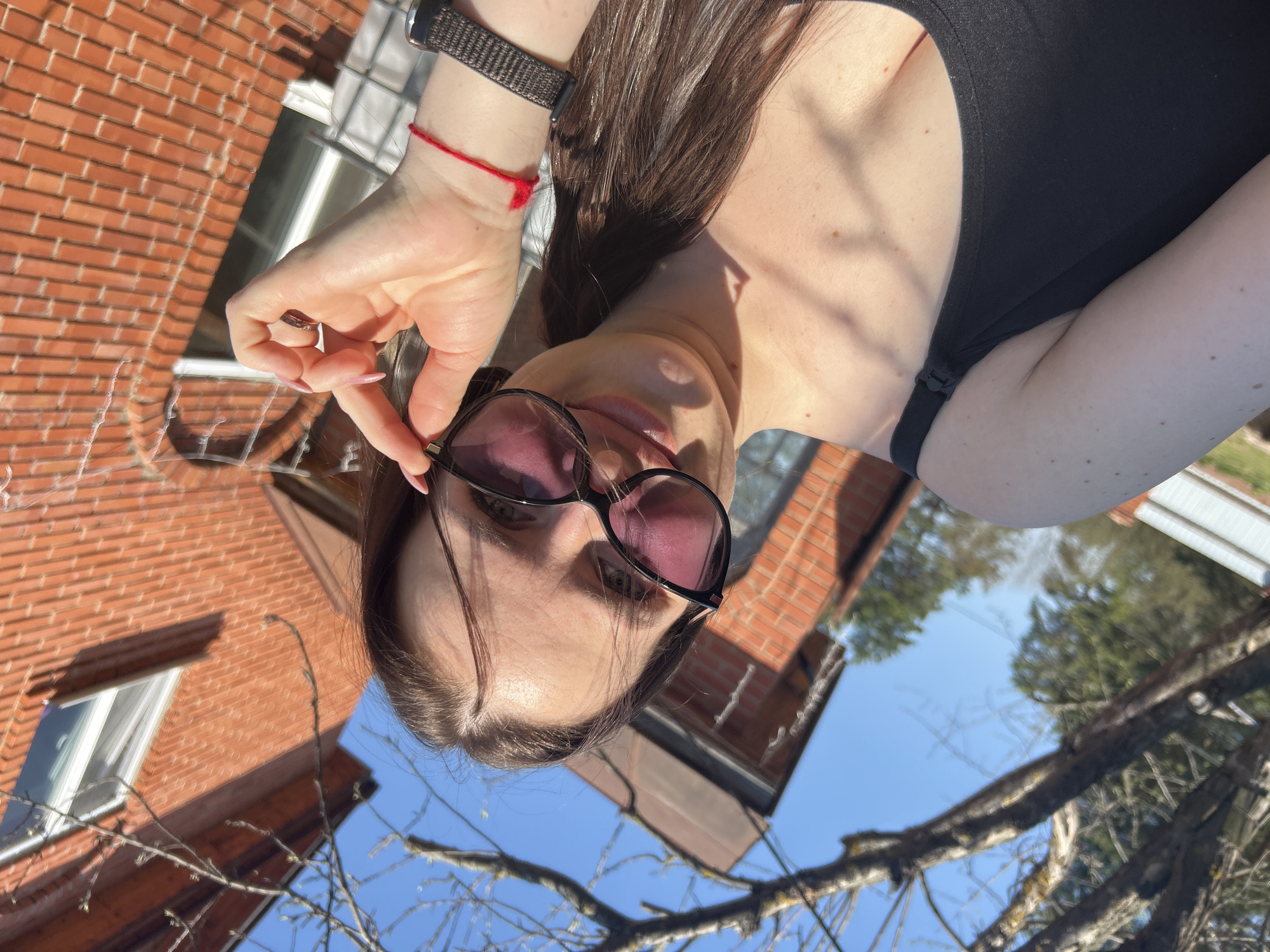 A woman with long, dark hair is taking a selfie outdoors. She is wearing sunglasses and a black top. A red string bracelet is visible on her wrist. In the background are bare tree branches against a bright blue sky and a brick building with windows. The location is Rusalevka, Russia.