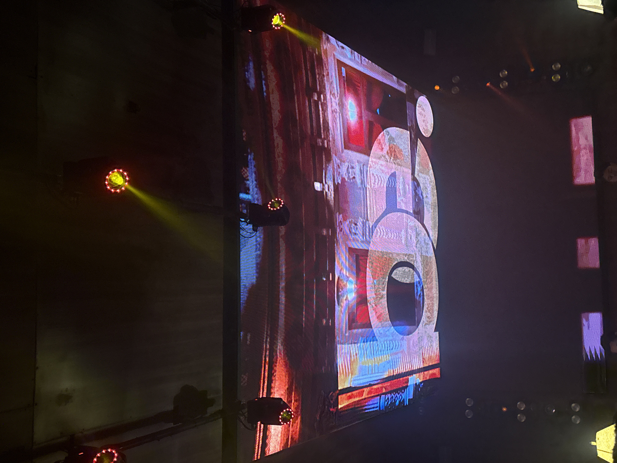 The image captures a large LED screen displaying a colorful graphic, possibly a stylized "60" or a similar design. The screen exhibits a mix of red, blue, and white hues, with prominent circular shapes within the graphic. The screen is flanked by stage lighting, with at least four spotlights visible, casting beams of light onto the screen and the surrounding area. The location appears to be a stage or concert venue, as evidenced by the lighting setup and the dark backdrop. The image does not provide enough information to confirm the location in Puebla, Mexico. There are no people visible in the frame, and the primary focus is on the screen and lighting.