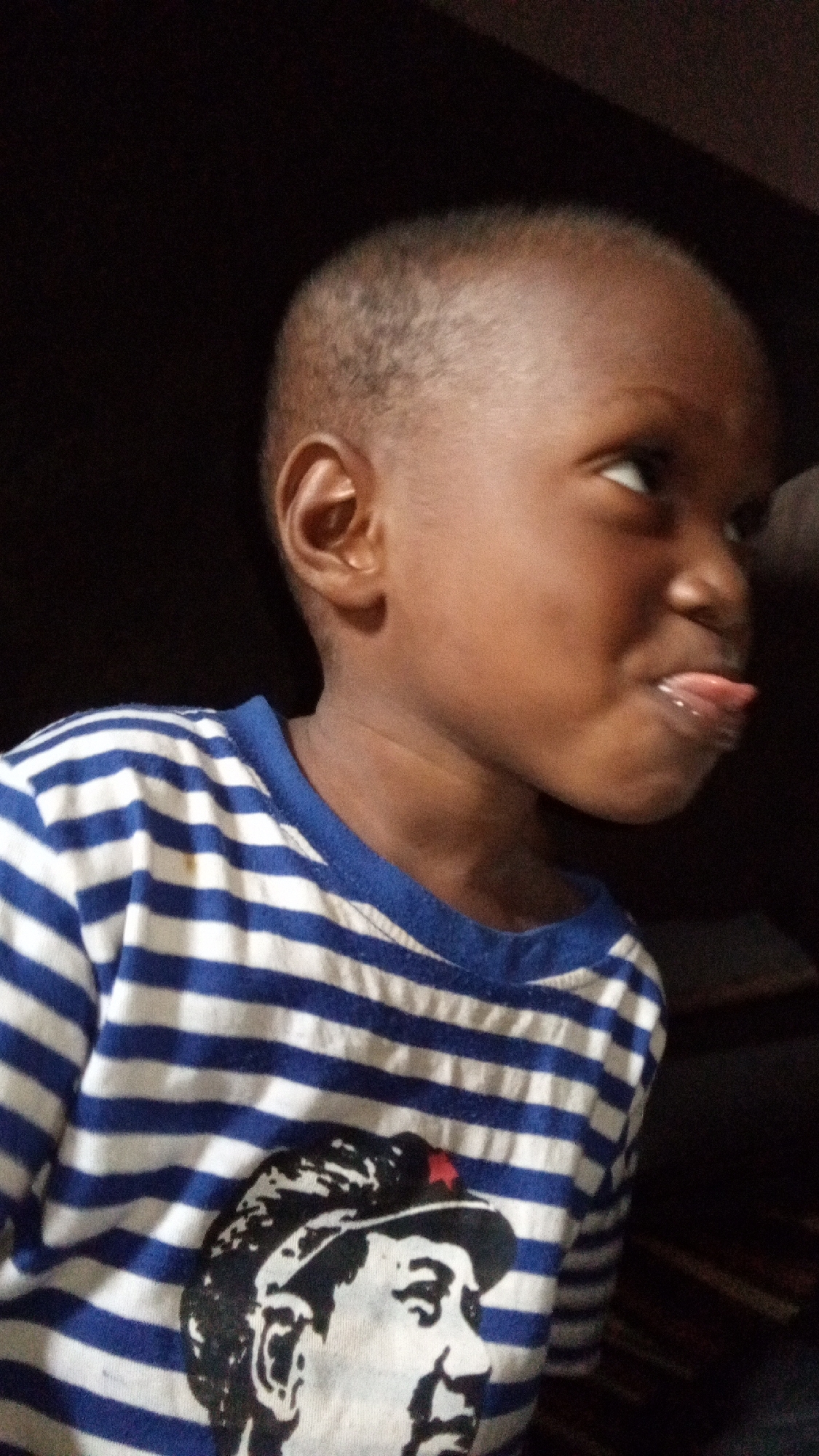A close-up shot of a young child, likely male, with dark skin and a short haircut, wearing a blue and white striped shirt. The shirt has a graphic of a person on it. The child is sticking their tongue out and looking to their right. The background is dark and indistinct. It is not possible to verify if this photo was taken in Sabon Tasha, Nigeria.