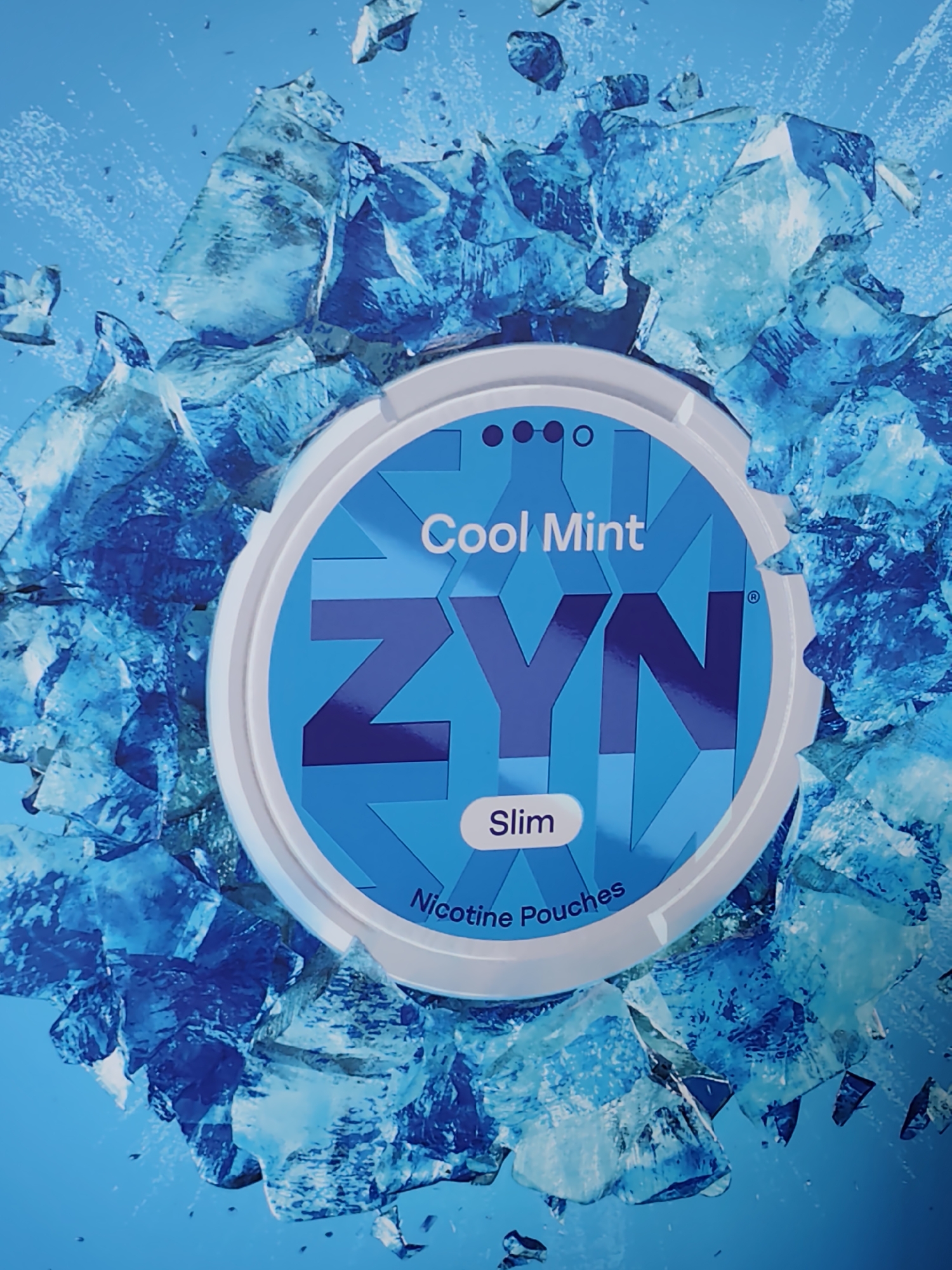 The media file is a close-up, eye-level product shot of a ZYN Cool Mint nicotine pouch container. The container is round with a white rim and a bright blue lid. The lid features the brand name "ZYN" in large, stylized dark blue letters, with a smaller "Cool Mint" label above it and "Slim" in an oval below. The text "Nicotine Pouches" is also visible near the bottom rim.
The container is presented against a vibrant blue background, which is visually fragmented by a dynamic arrangement of shattered ice or crystal-like shapes. These icy elements appear to be exploding outwards from behind the container, creating a sense of motion and a cool, refreshing aesthetic. The ice fragments are various shades of blue, from deep indigo to pale cyan, with sharp, angular edges and some appearing translucent. Small water droplets or ice shards are scattered in the air around the main ice formations.
There are no people visible in the image. The focus is solely on the product and the thematic icy background. The lighting appears bright and even, highlighting the details of the product and the frosty elements. The overall mood conveyed is one of extreme coolness and a crisp, invigorating sensation, aligning with the "Cool Mint" branding. The setting is abstract and studio-like, designed to emphasize the product's flavor profile.