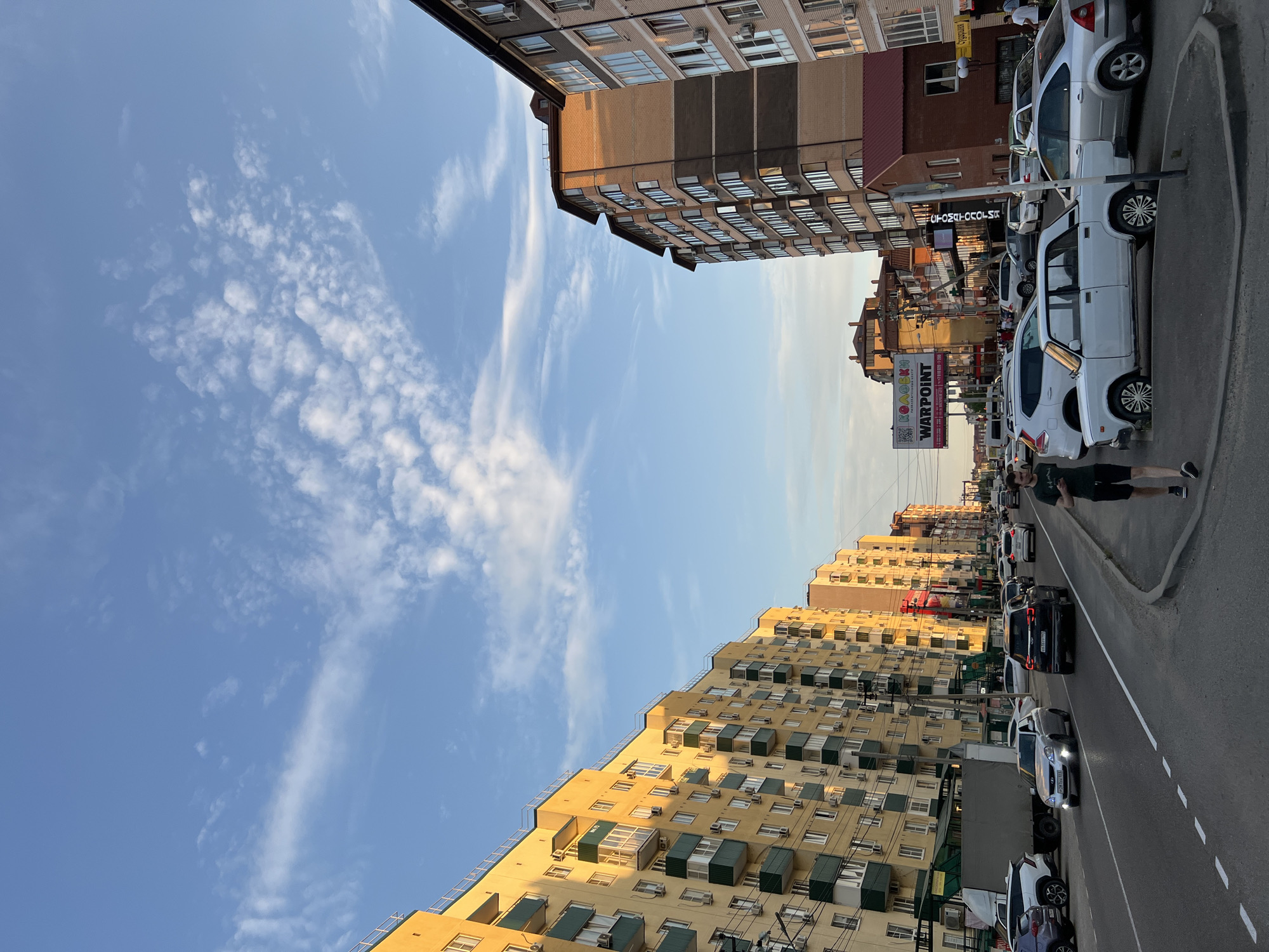 The image depicts a street scene in Yablonovskiy, Russia, characterized by apartment buildings and a busy road. The sky above is blue with scattered white clouds, suggesting it is either morning or late afternoon.
On the left side of the street, a long, multi-story apartment building with a yellow facade and green balconies dominates the view. The building appears to be residential, with numerous windows and air conditioning units visible.
On the right side of the street, several modern apartment buildings are visible. The closest one has a brick facade with dark horizontal bands and numerous windows. Further down the street, other buildings with light-colored facades are present.
The road is paved and appears to be a main thoroughfare, with multiple lanes of traffic. Several cars are driving on the road, including a silver sedan, a dark SUV, and a white sedan. A large, grey truck is parked on the left side of the road.
In the foreground on the right, a man is walking along the sidewalk, looking at his phone. He is wearing a dark t-shirt and shorts. The sidewalk is bordered by a curb and a strip of grass.
A billboard with the text "WARPOINT" is visible in the distance, along with other signs and storefronts, indicating commercial activity in the area. The overall impression is of an urban environment with residential and commercial elements, bustling with daily activity.