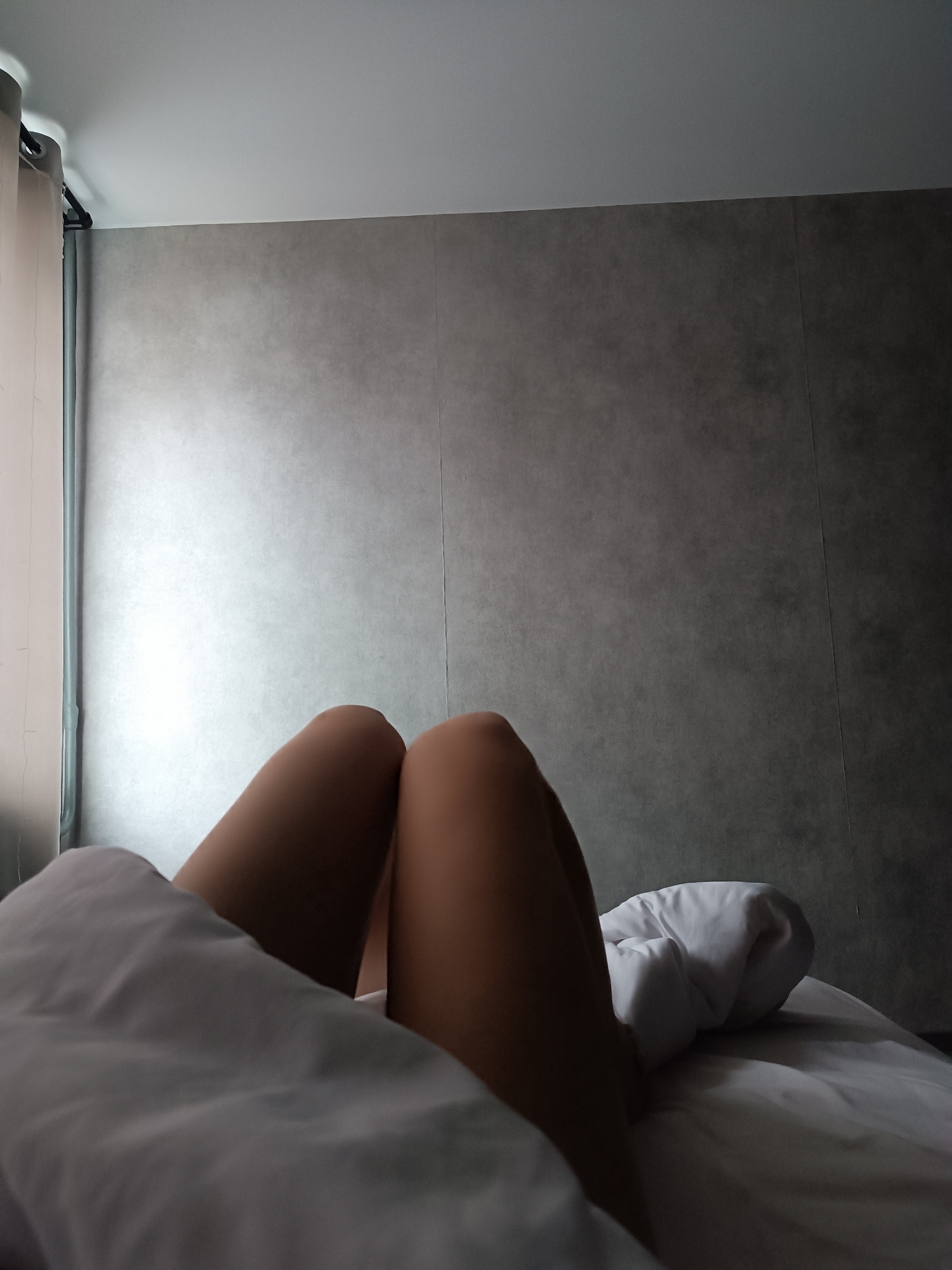 The image shows a person lying in bed, with their legs exposed from beneath a white blanket. The room has a grey wall and a window with a curtain on the left side. The location is unknown.