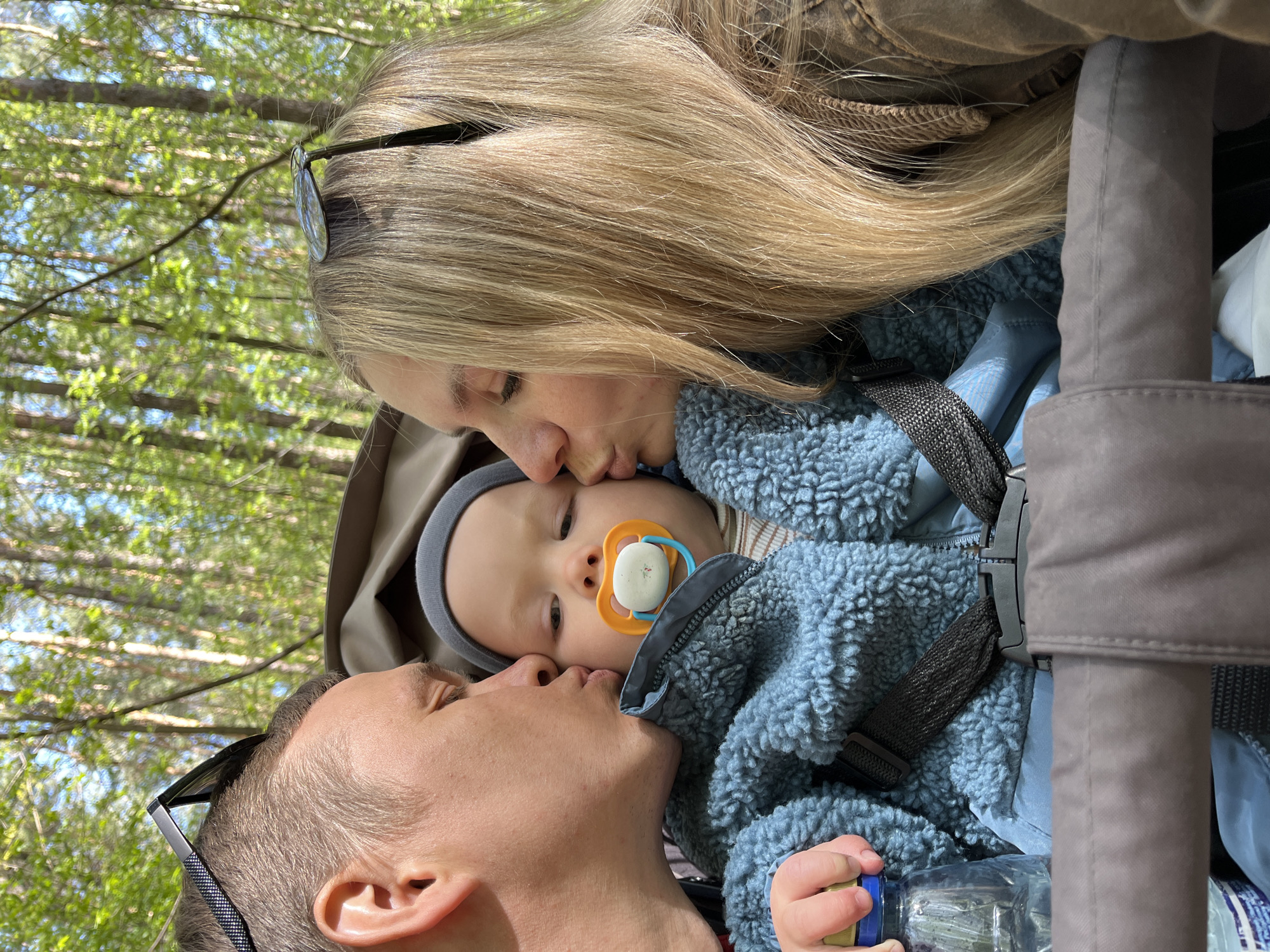 A close-up shot captures a family moment, likely outdoors given the blurred trees in the background. A blonde woman with sunglasses perched on her head is kissing a baby’s forehead. The baby, wearing a grey hat and a blue fleece jacket, has a pacifier in its mouth. A man with closely-cropped hair and sunglasses also perched on his head, kisses the baby's cheek. The baby is strapped into a carrier, holding a plastic water bottle. The location is not discernible from this image.