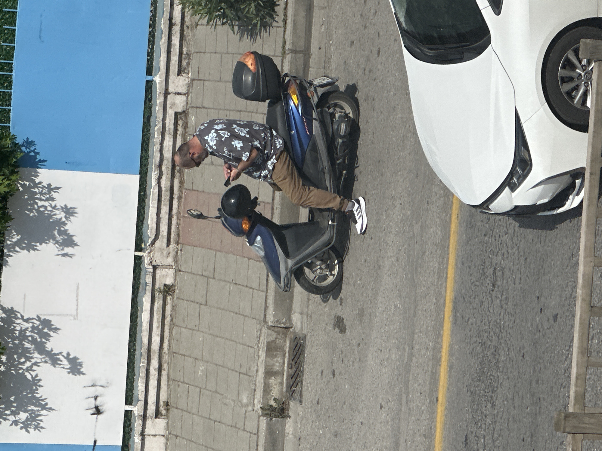 A man in a floral shirt and tan pants is standing next to a blue and black scooter on a street. The scooter is parked on the side of the road next to a sidewalk paved with gray bricks. A white car is parked adjacent to the scooter, partially in the road. A yellow line marks the center of the road. A building is visible to the left of the frame with a white wall and blue upper section. The scene is located in an urban area with trees and vegetation. Based on the architecture and vegetation, the location is likely Fethiye, Turkey.