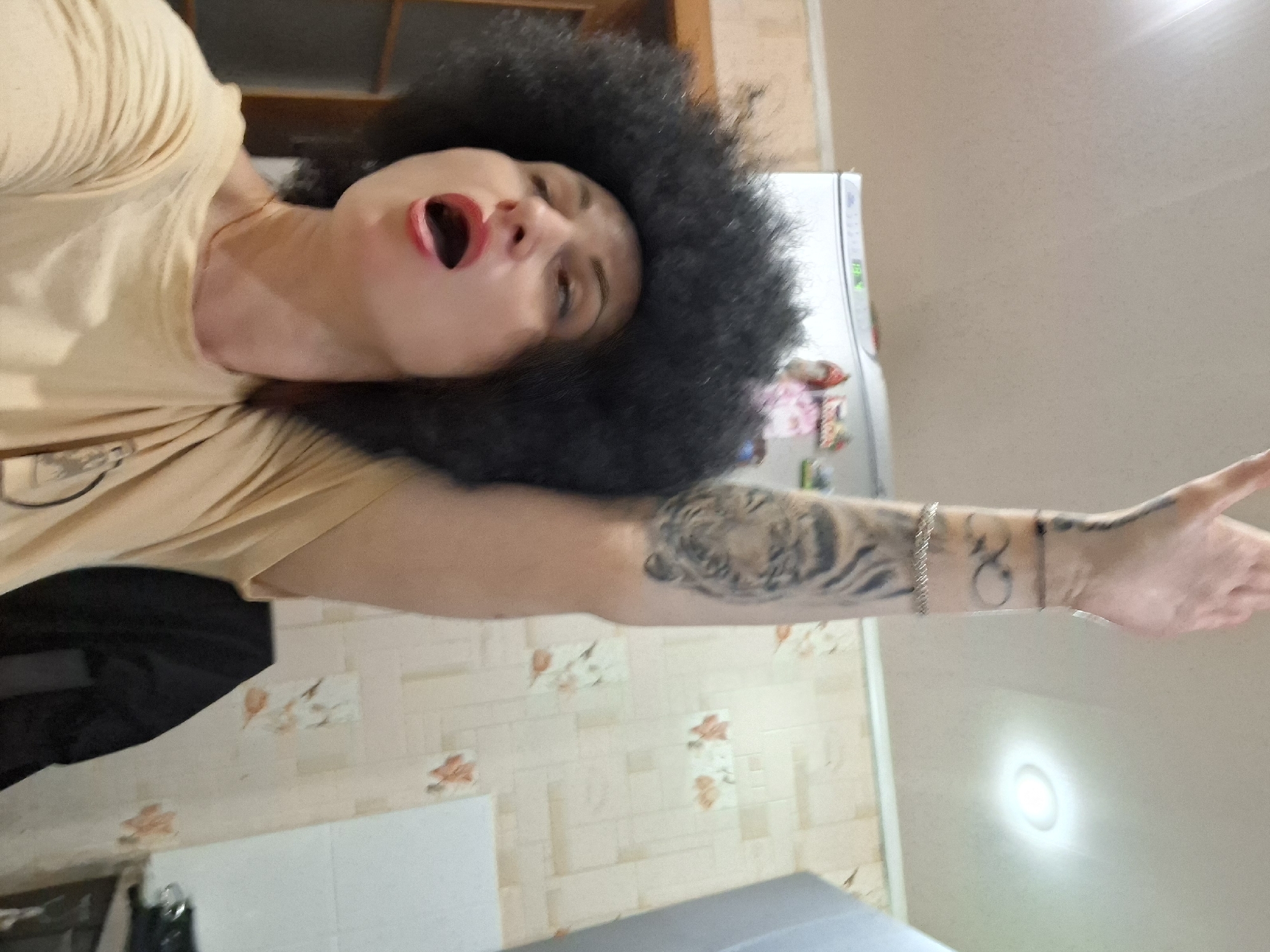 A woman with an afro wig and an open mouth is seen with her right arm raised upwards in a dimly lit indoor environment, possibly a kitchen, given the visible refrigerator and tiled walls. The woman is wearing a light-colored t-shirt. She has tattoos on her arm, including what appears to be a tiger's face and an infinity symbol. The overall lighting is soft, with a ceiling light source visible. The background consists of light-colored, patterned wallpaper and a white refrigerator. The scene is a close-up shot, focusing on the woman and her arm. There is no indication of the city of Myadzyel, Belarus.