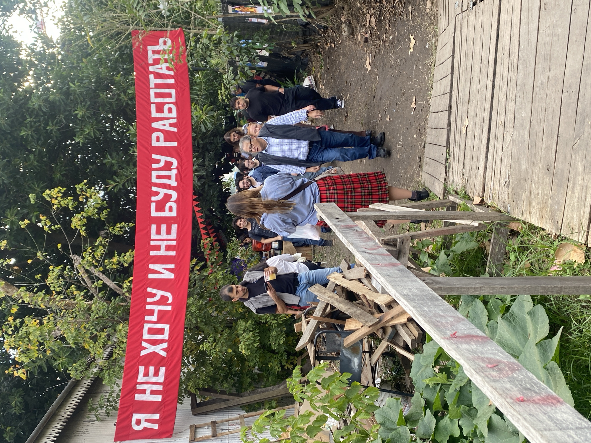 A photograph captures a group of people gathered outdoors on a dirt and wood-plank path amidst lush greenery. A striking red banner, positioned vertically, displays a phrase in white Cyrillic letters. People are wearing a variety of clothing including blue jeans, dresses, and shirts. The scene is illuminated by natural daylight, suggesting an outdoor setting in the city of Buenos Aires, Argentina.
