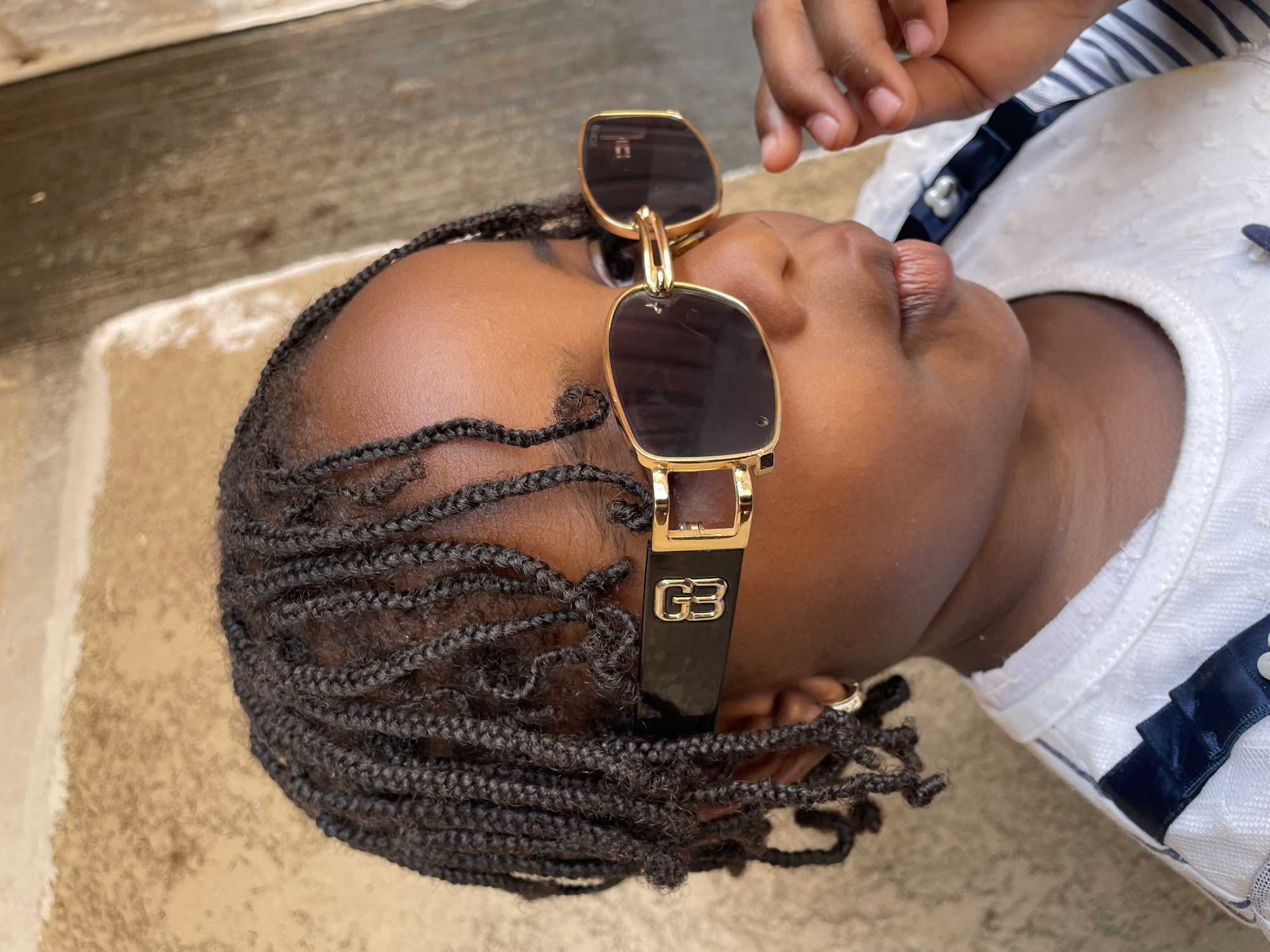 A close-up, high-angle photograph shows a child with dark skin, presumably in Jos, Nigeria, wearing sunglasses perched on their head. The child has braided hair. They are wearing a white shirt with a dark blue and white striped collar. The child's hand is partially visible above their face. The background appears to be a concrete or stone surface.