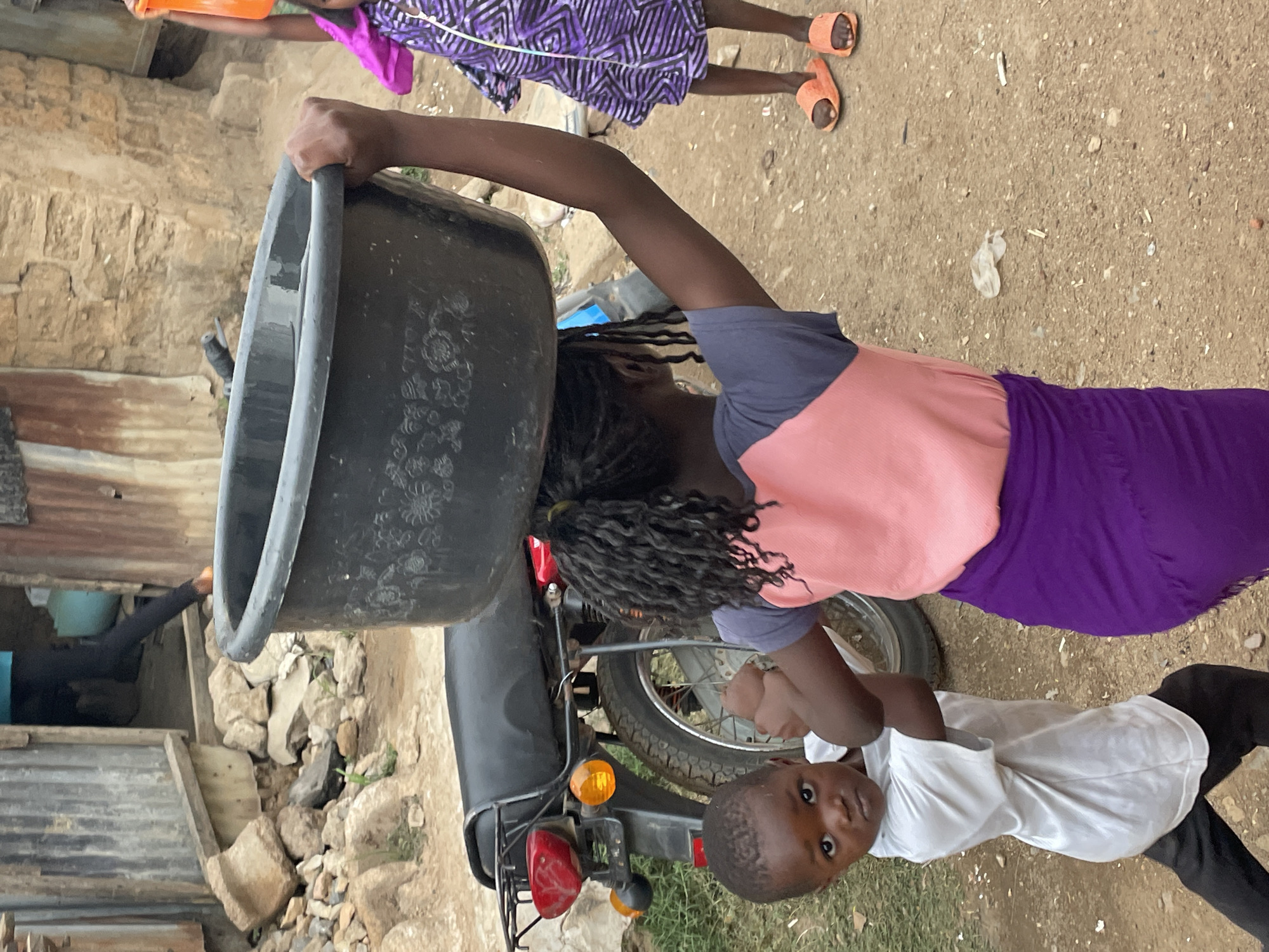 The image depicts an outdoor scene in Jos, Nigeria, featuring three children. In the foreground, a young girl is balancing a large, round black plastic basin on her head, her face partially obscured by it. She wears a peach-colored top, a purple skirt, and has dark, braided hair or dreadlocks falling down her back. Her right hand is raised, seemingly steadying the basin.
To her right, a younger boy in a white t-shirt and black trousers stands, looking directly at the camera with a neutral expression. Behind the girl, a black motorcycle is parked, with its rear wheel, fender, and tail lights (red and orange) visible. Further in the background, another child, wearing a purple and white patterned dress and orange slip-on sandals, is standing.
The ground is a mix of dirt and small stones, indicative of a rugged outdoor setting. The background features rudimentary structures, including walls made of rough-hewn stone blocks and sections of corrugated metal, suggesting a modest residential or semi-urban environment. The lighting is natural and appears to be daytime, possibly overcast. The basin itself has a subtle embossed floral design on its side. There is no visible text in the image.