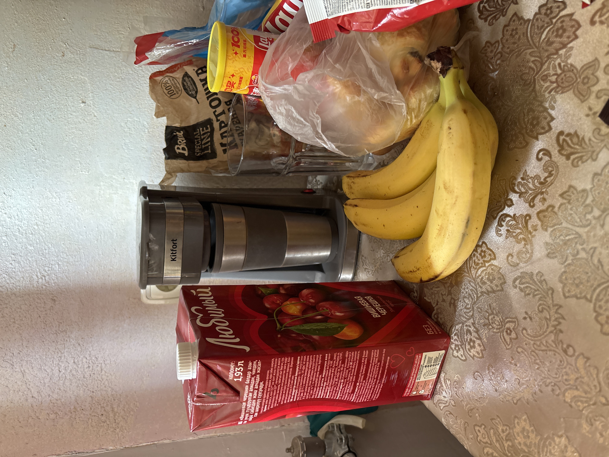 This is a still life photograph taken indoors, likely in a kitchen or dining area, with a close-up view of various food items and kitchen appliances on a table. The background consists of a textured, off-white wall.
In the foreground and middle ground, the following objects are visible:
* A large, red carton of "Любимый" (Lyubimyy) cherry juice, with text in Russian.
* A bunch of ripe bananas, with three or four individual bananas prominently displayed.
* A clear glass.
* A plastic bag containing bread or pastries.
* A brown paper bag labeled "Brik Special Line" with Russian text, possibly containing potato products.
* A yellow can with red Chinese characters and "100%" text.
* A red and white bag, partially visible, with similar Chinese characters and "100%" text.
* A red packet with white text.
* A coffee maker with a "Kitfort" logo.
* A metal faucet fixture on the far left, partially obscured.
The scene is well-lit, with shadows cast by the objects onto the patterned tablecloth and the wall. The overall impression is of a domestic setting where groceries are unpacked or displayed. There are no people or animals present.
There is no specific information within the image that indicates the location in the city of Saint Petersburg, Russia.