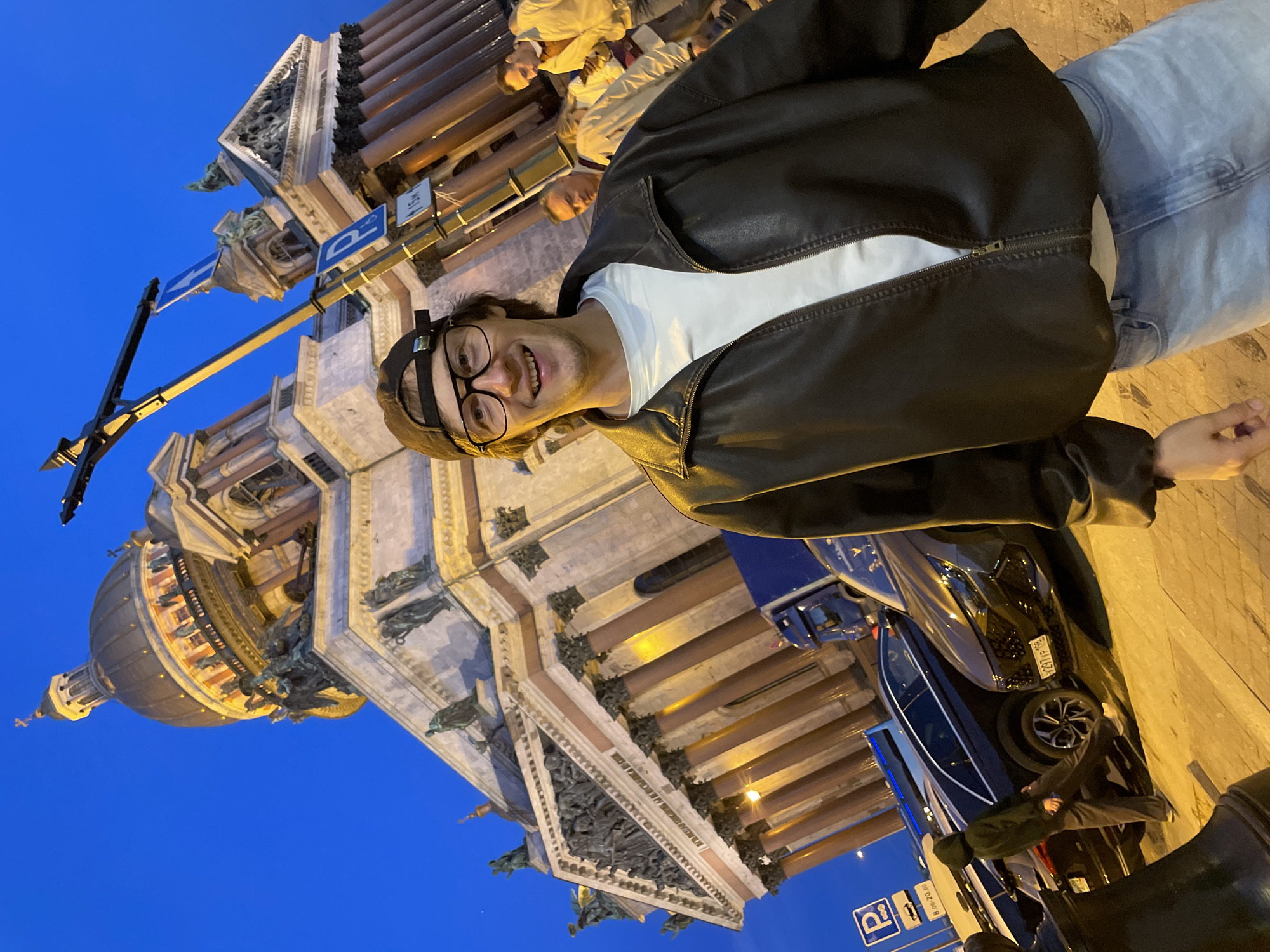 The image shows a young man wearing a black leather jacket, a white t-shirt, blue jeans, glasses, and a baseball cap, smiling at the camera. Behind him, St. Isaac's Cathedral, a prominent landmark in Saint Petersburg, Russia, is visible. The cathedral's dome and facade are illuminated against a twilight sky. In the foreground, several cars are parked, and some people are walking in the background. The scene appears to be a candid photograph taken outdoors at dusk.