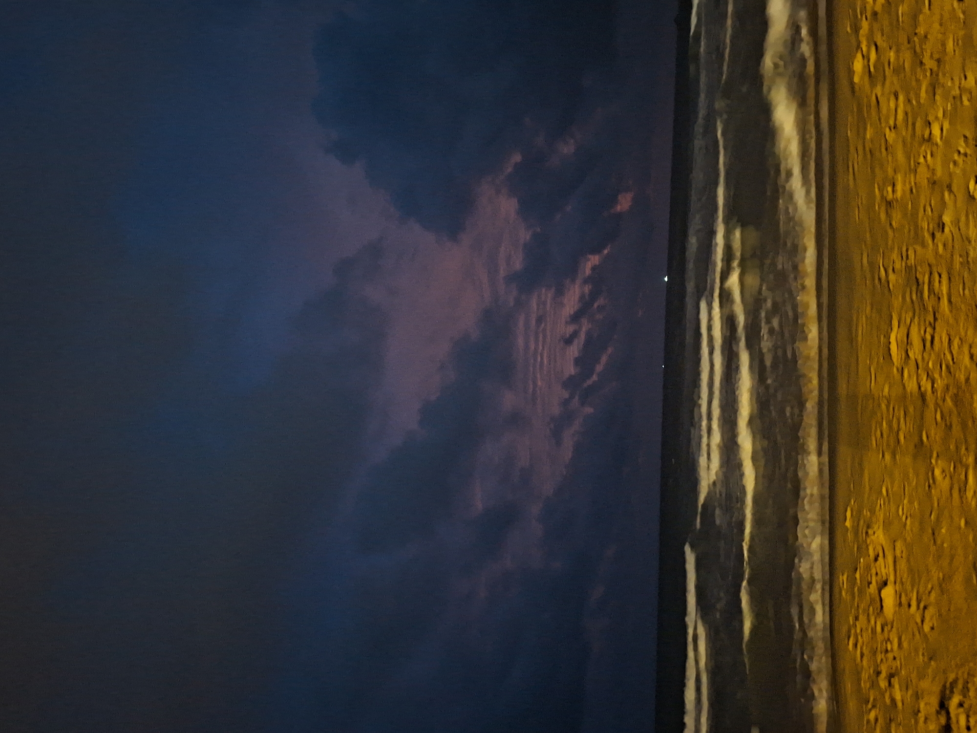 The image depicts a beach scene at dusk in Phu Quoc, Vietnam. The sky is a deep blue, transitioning to hues of purple and pink near the horizon, with dark, cloudy formations dominating the upper portion of the frame. The sea is calm, with gentle waves rolling towards the shore, creating streaks of white foam against the dark water. The foreground is a sandy beach, illuminated by an artificial light source, casting a warm, golden glow on the textured sand. There are no people or discernable objects on the beach or in the water, except for what appear to be distant lights on the horizon, possibly from boats or coastal structures. The overall impression is one of a tranquil, natural landscape at the end of the day.