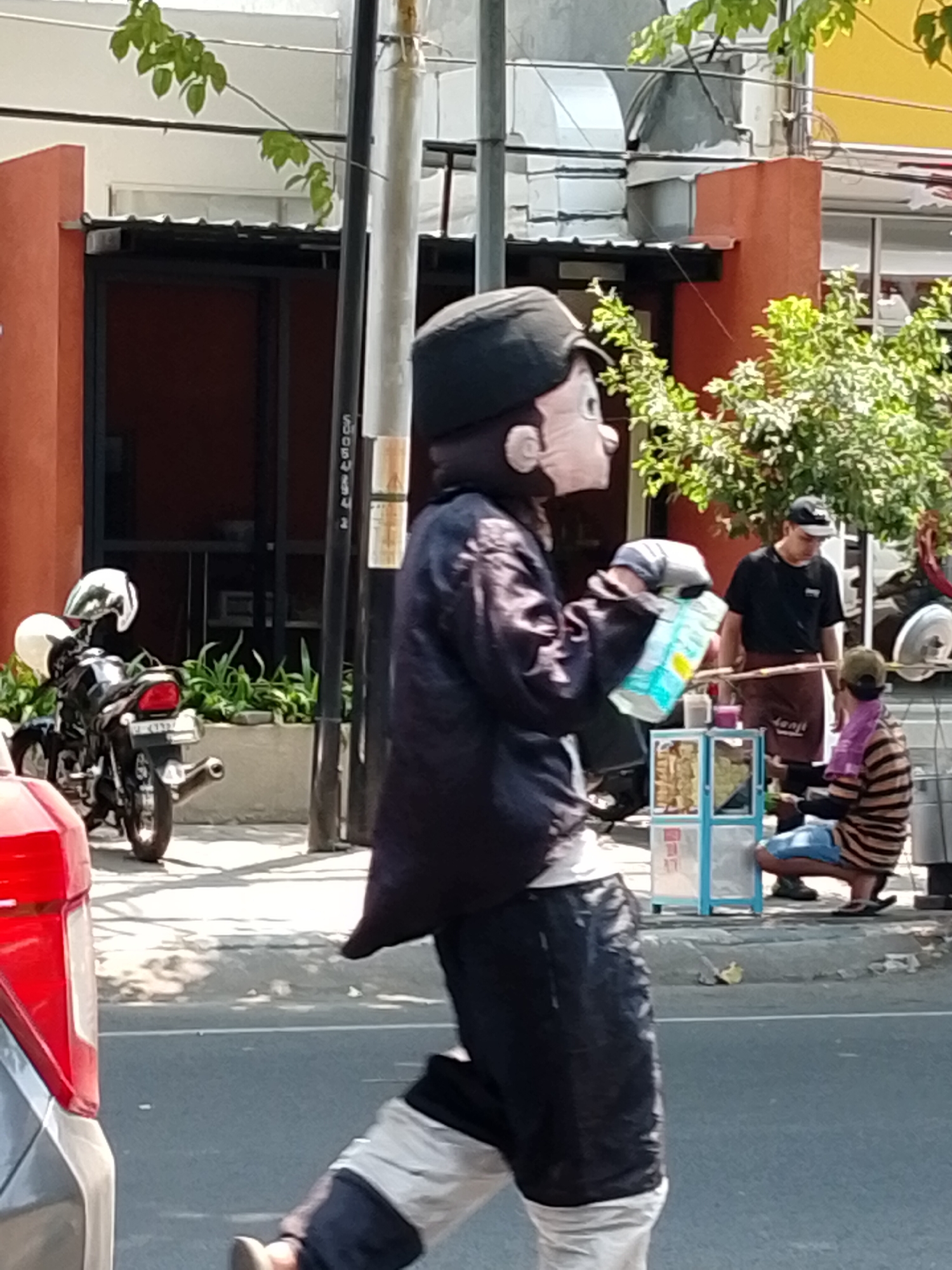 A person in a costume that resembles a cartoon character is walking on a sidewalk in Pasuruan, Indonesia. The costume has a large, pale face with round cheeks, a prominent nose, and dark-colored hair. They are wearing a dark, satin-like jacket over a white undershirt and dark pants. The character is holding a light blue and white box or package in their right hand.
In the background, a black motorcycle with two white helmets is parked. A man wearing a black t-shirt and a brown apron stands behind a food cart. To the right of the food cart, a person wearing a striped shirt and blue shorts is crouching. The environment appears to be a street with buildings and trees visible. The lighting suggests it is daytime.