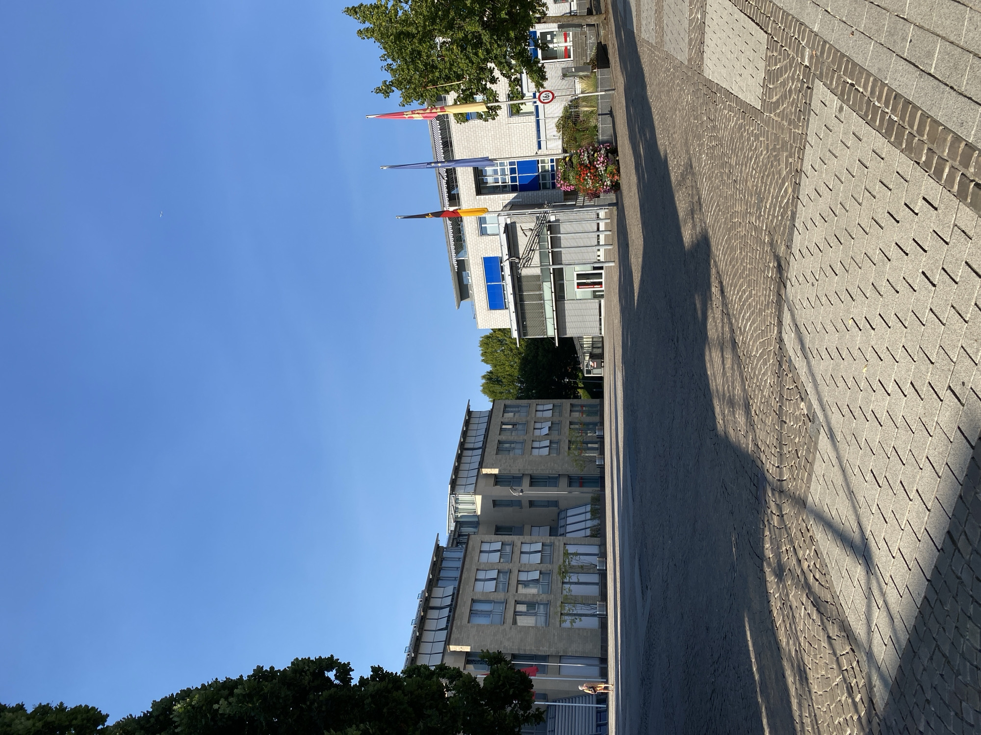 This media file depicts a sunny day in Altenkirchen, Germany. The scene captures a modern architectural setting with two multi-story buildings. The building in the foreground, on the right side of the frame, is clad in gray stone and features numerous windows, many of which are open. It has a sleek, contemporary design with balconies and glass facades. The building behind it, partially obscured by trees, appears to be made of lighter-colored brick and also has a modern design with prominent blue window frames.
In front of the brick building, several flags are visible, including what appears to be the German flag and possibly the European Union flag. Colorful flowers are arranged in planters near the entrance, adding a touch of vibrancy. A speed limit sign with "30" is also visible, indicating a 30 km/h zone. The ground is paved with cobblestones in intricate patterns, leading away from the buildings and casting long shadows, suggesting it is mid-to-late afternoon.
A lone person is visible near the entrance of the gray stone building, appearing to walk away from the camera. The sky is a clear, bright blue, and the presence of lush green trees indicates a pleasant, likely summer, day. The overall impression is one of a well-maintained urban or civic space, bathed in warm sunlight.