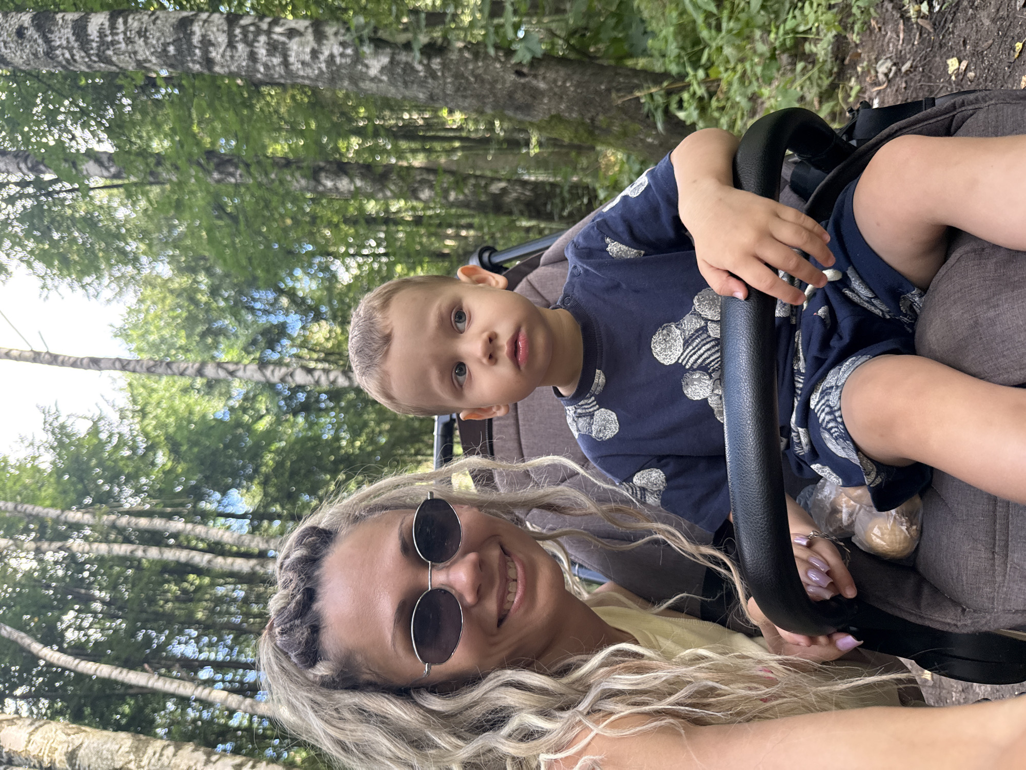 In Rusanovo, Russia, a woman with blonde curly hair and sunglasses smiles at the camera while sitting next to a young boy in a stroller. The boy, wearing a dark blue t-shirt with a circular pattern, looks to his right with a neutral expression. The scene is set in a forest with birch trees and lush green foliage visible in the background. A stroller handle separates the woman and the child, and a plastic-wrapped object, possibly food, rests in the stroller.