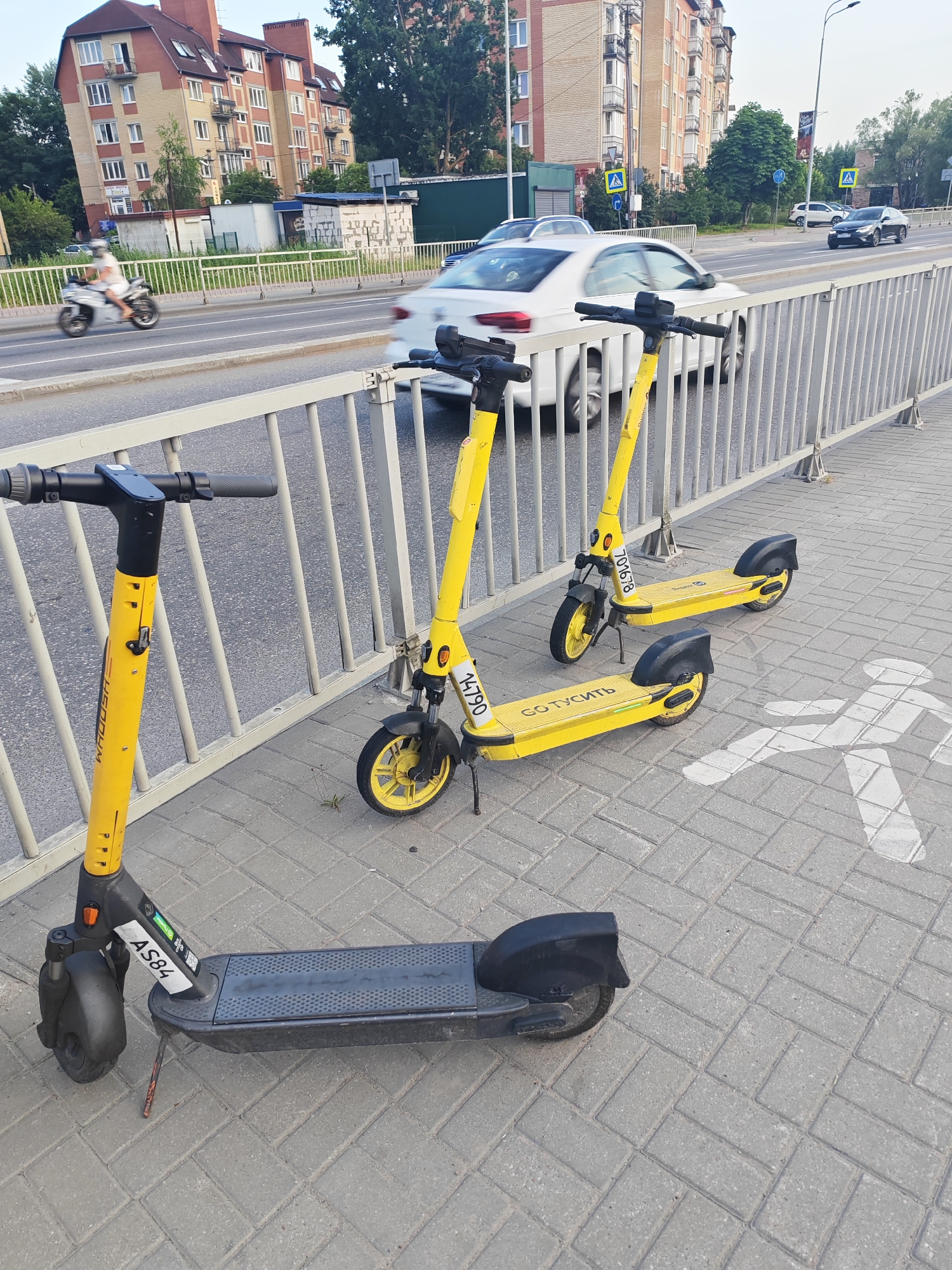 In the city of Vasilkovo, Russia, a sidewalk is lined with three yellow rental electric scooters. The scooters are parked parallel to a metal fence and a street with cars. The pavement is made of light gray interlocking bricks. A white silhouette of a person walking is painted on the pavement to the right of the scooters, indicating a pedestrian crossing. In the background, a multi-story brick building with red-tiled roofs and balconies can be seen. Trees and other buildings are also visible along the street. The sky is overcast.