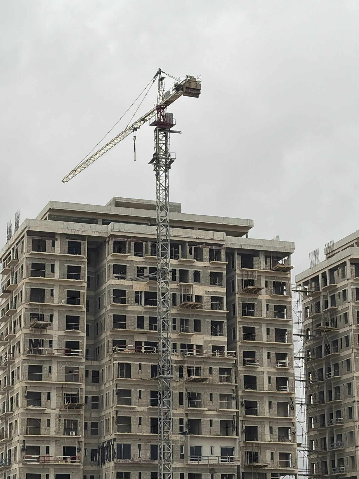 An overcast sky hangs above a construction site in Alaguntan, Nigeria, where two partially completed multi-story buildings dominate the frame. A tall construction crane, its yellow boom extended diagonally across the sky, is positioned between the two structures. The buildings themselves are bare concrete skeletons, featuring rows of dark, empty window openings and exposed rebar at their tops. Construction scaffolding is visible on the exterior of the buildings. The scene is devoid of people, suggesting ongoing work or a pause in activity.