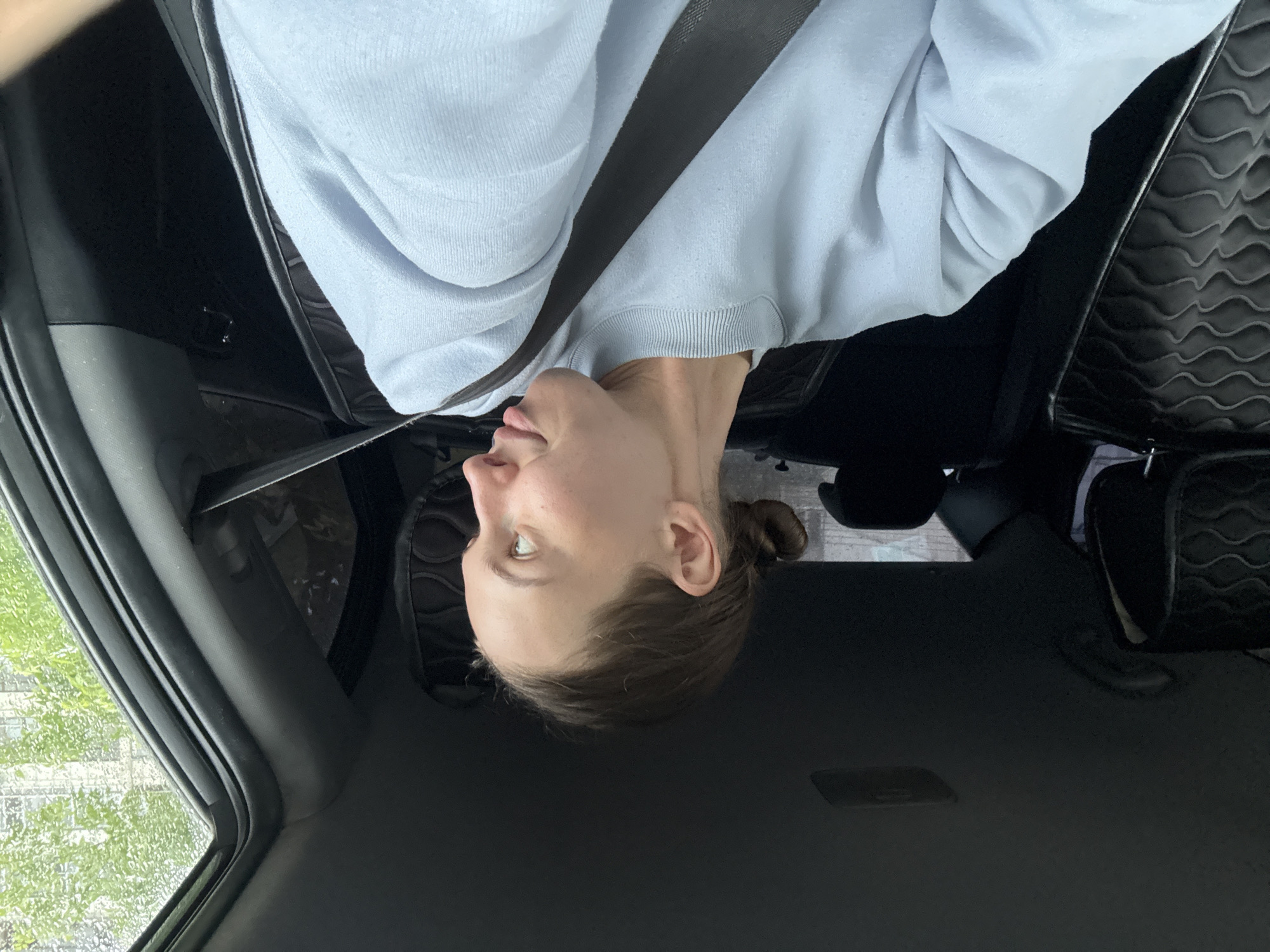 A woman is seated in the passenger seat of a car, looking over her right shoulder towards the outside of the vehicle. She has her hair pulled back in a bun and is wearing a light blue sweatshirt and a seatbelt. The interior of the car is visible, including the headrests and seats, and the window shows a blurred view of trees and buildings. The image appears to have been taken in the city of Perm, Russia.