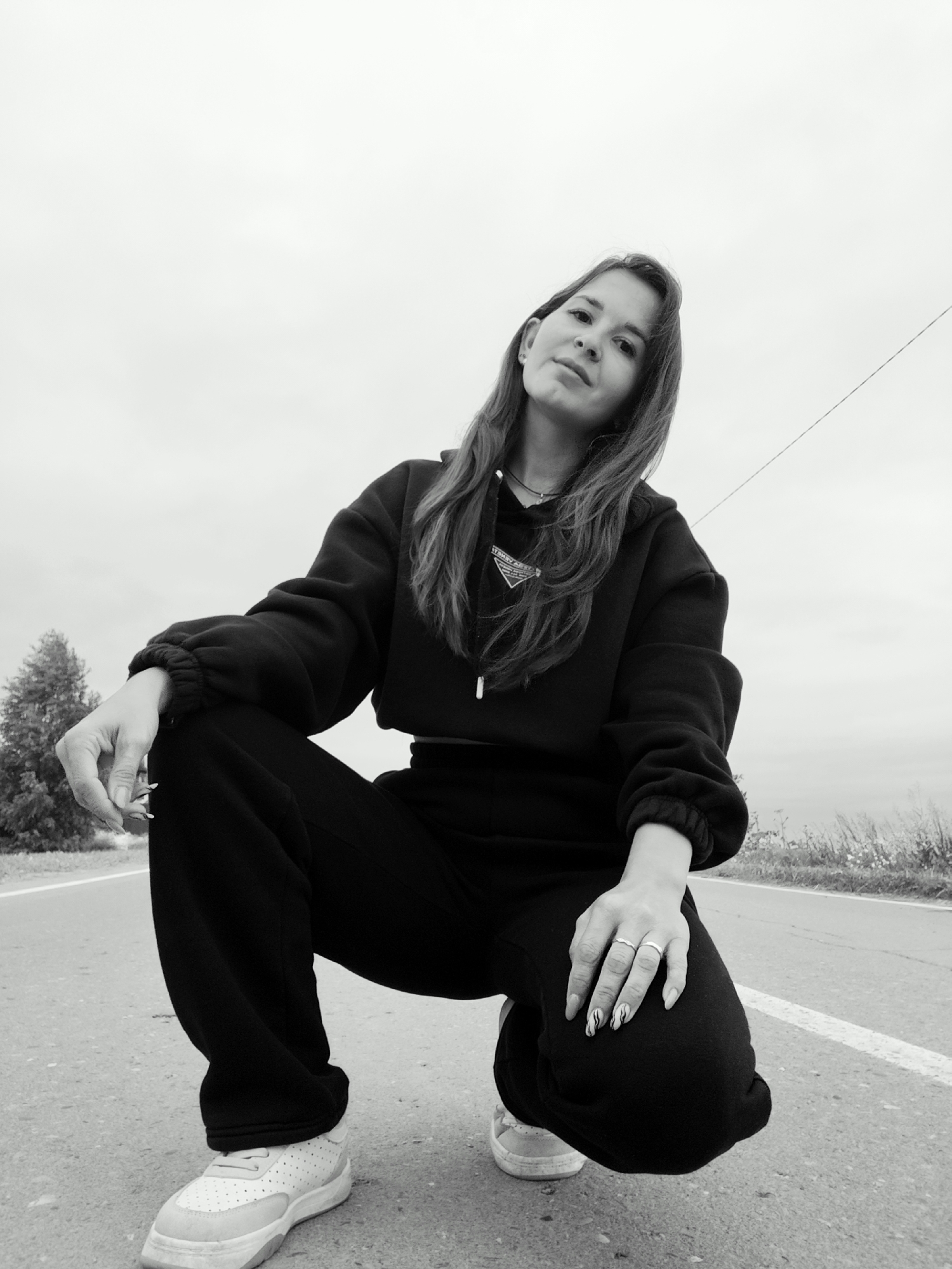 In a black and white photograph, a young woman is captured in a low-angle shot, crouching on an asphalt road. She is wearing a dark hooded sweatshirt and loose-fitting pants, with white sneakers on her feet. Her long, dark hair cascades around her shoulders, and she is wearing rings on her fingers. The background suggests an outdoor setting, possibly on the outskirts of a city, with a tree visible on the left and a thin power line stretching across the overcast sky. The road has a white line marking its edge, and sparse vegetation grows along its side. There is no direct evidence to confirm the location as Фоки, Russia, but the scene depicts a person in a rural or semi-urban environment.