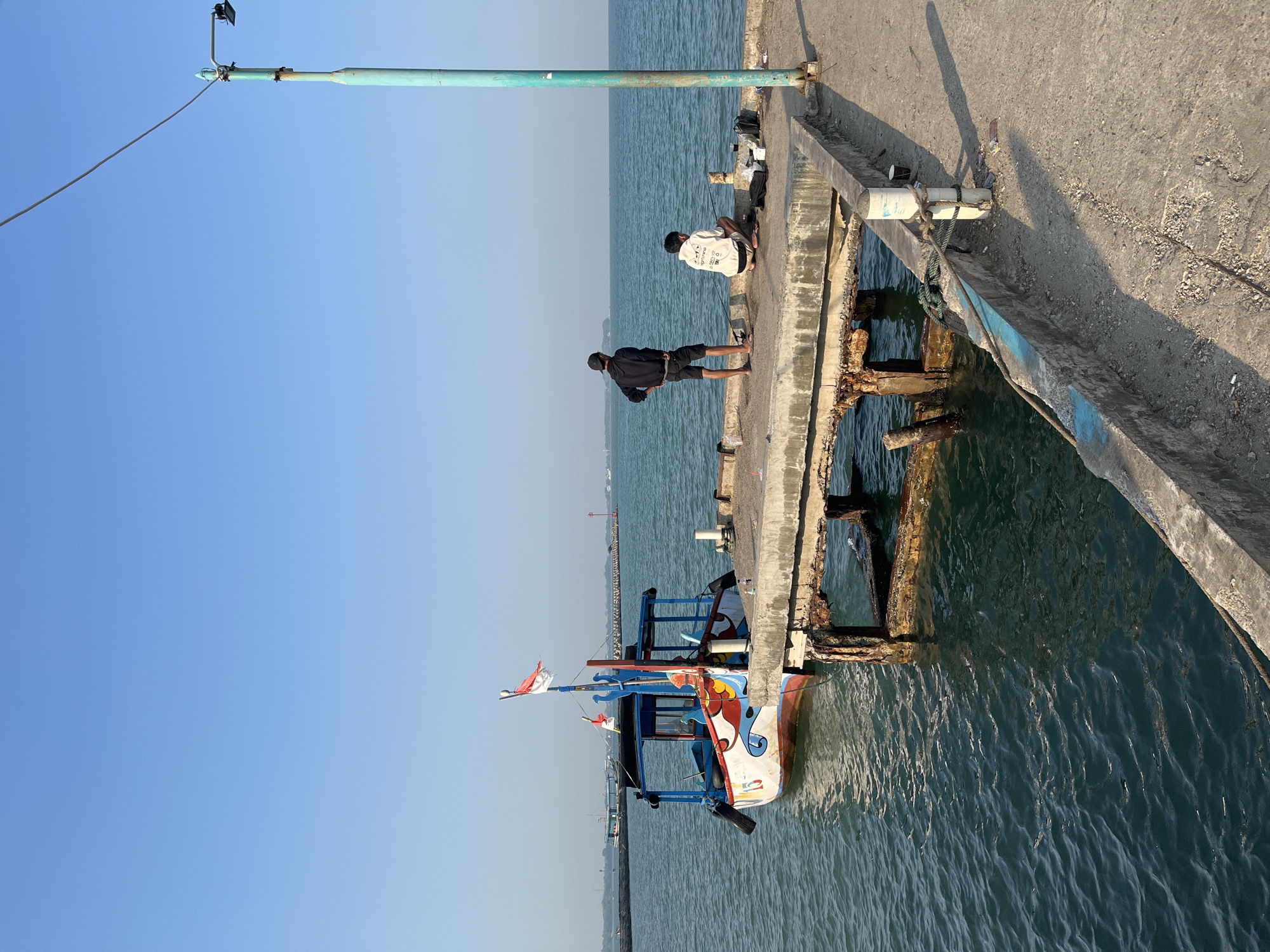 The image depicts a seaside scene in Jepara, Indonesia. In the foreground, a concrete pier extends into the ocean. A colorful, small fishing boat with a red and white Indonesian flag is moored to the pier. Two young men are present on the pier, one standing and observing the water, the other crouched down, possibly fishing or attending to equipment. The water is calm and blue, reflecting the clear sky. In the background, a breakwater or sea wall is visible, along with a distant lighthouse. A tall, teal-colored utility pole stands on the right side of the pier, with a wire extending across the frame. The overall atmosphere is serene, suggesting a peaceful day by the coast.