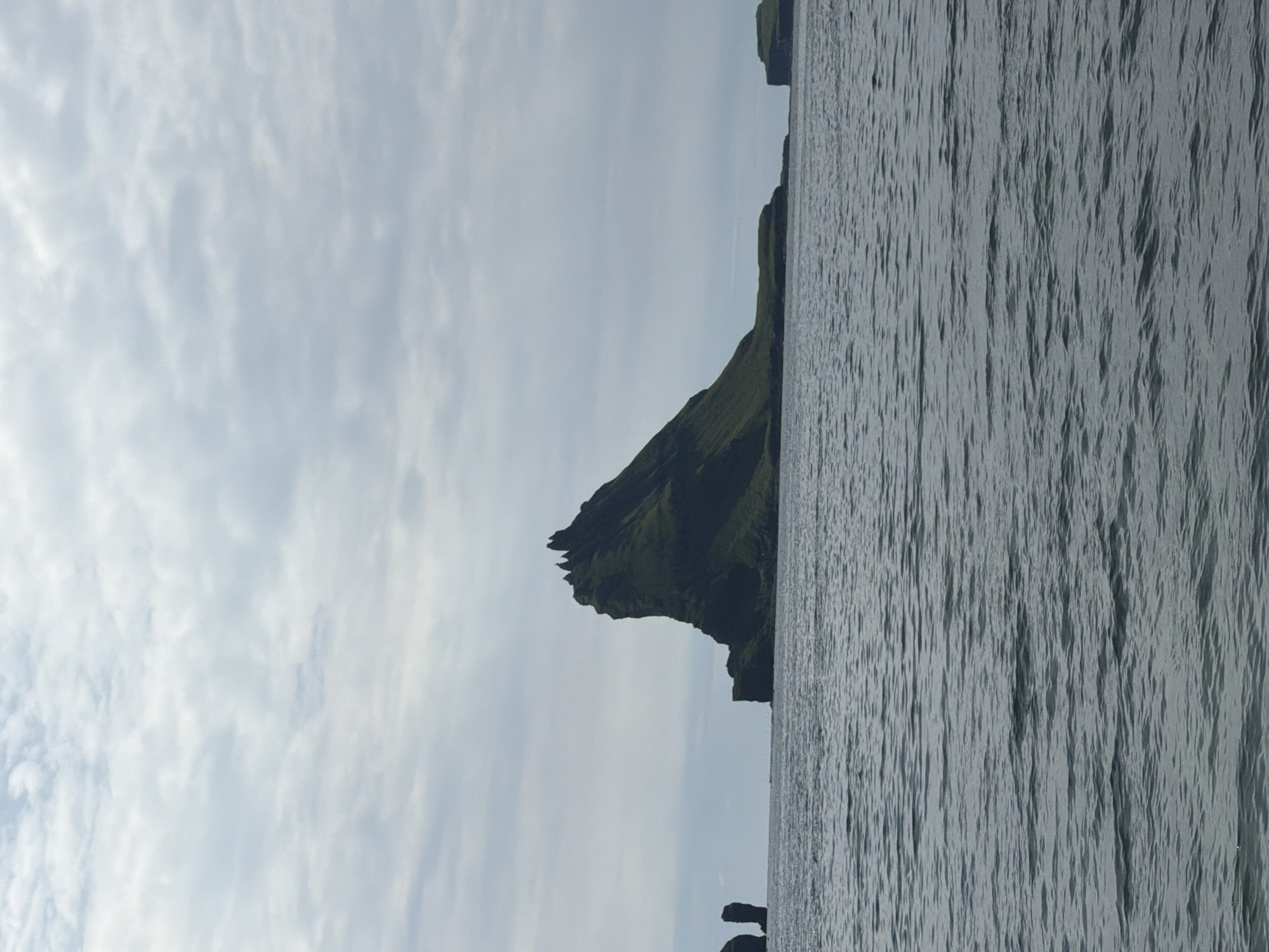 The image depicts a scenic view of the coast near Bøur, Faroe Islands. The foreground is dominated by the sea, with gentle waves creating a textured surface. In the midground, a prominent, dark green, jagged rock formation rises dramatically from the water, its steep slopes covered in vegetation. To the left of this main rock, a smaller, columnar rock formation stands in the sea. Further to the right, a lower, flatter island with grassy slopes is visible. The background consists of a cloudy sky, with soft, diffused light indicating an overcast day. The overall impression is one of natural beauty and rugged coastal landscape.