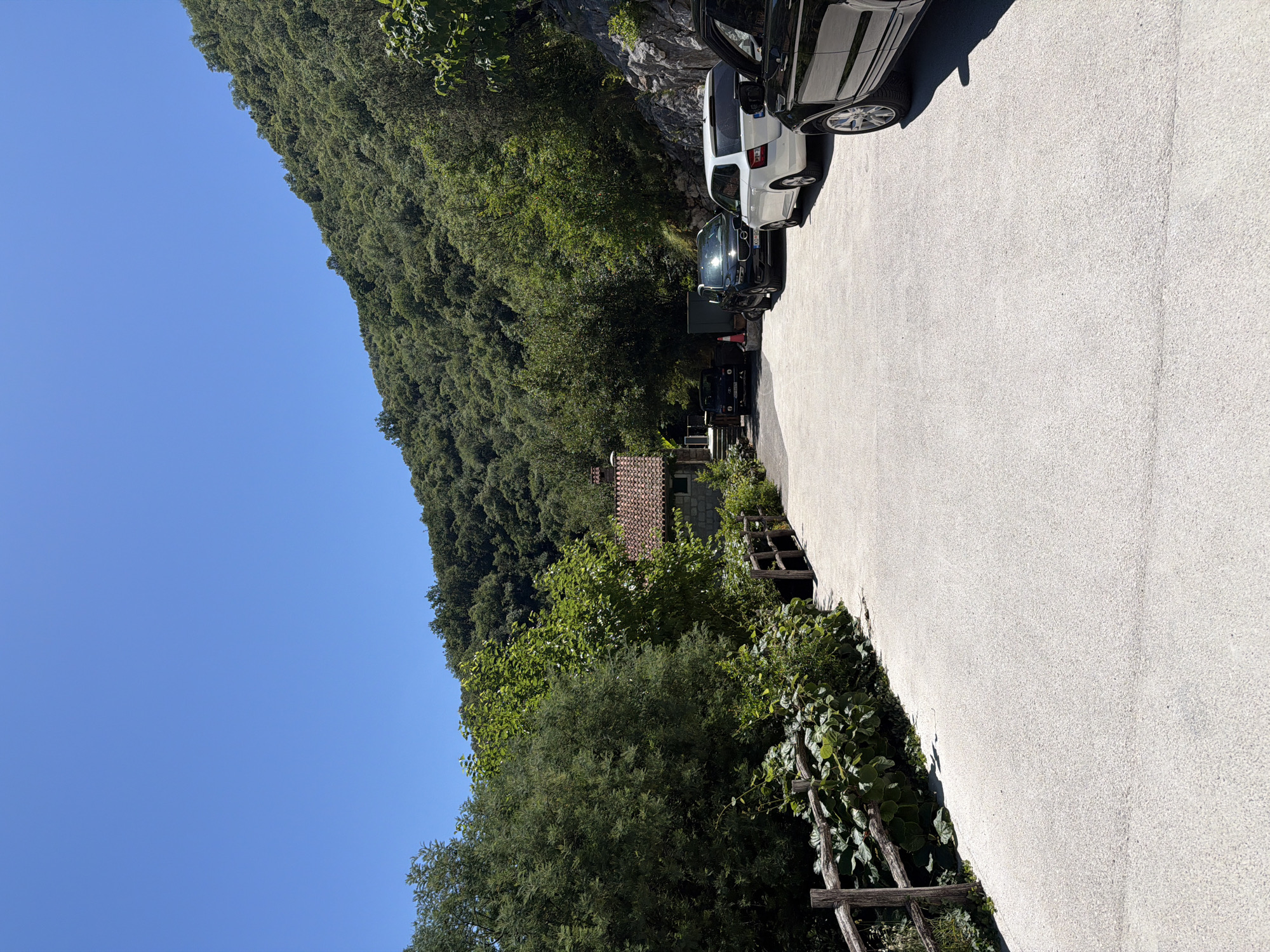 In the city of Morinj, Montenegro, a paved parking area is situated in a natural environment. The foreground is dominated by the light-colored asphalt of the parking lot, with a weathered wooden fence and lush green foliage lining the left side. In the background, a small building with a red tile roof is nestled amongst dense trees. Several vehicles are parked on the right side of the lot: a black SUV, a white hatchback, and another black vehicle partially visible. The scene is set against a backdrop of a steep, tree-covered hillside under a clear blue sky.