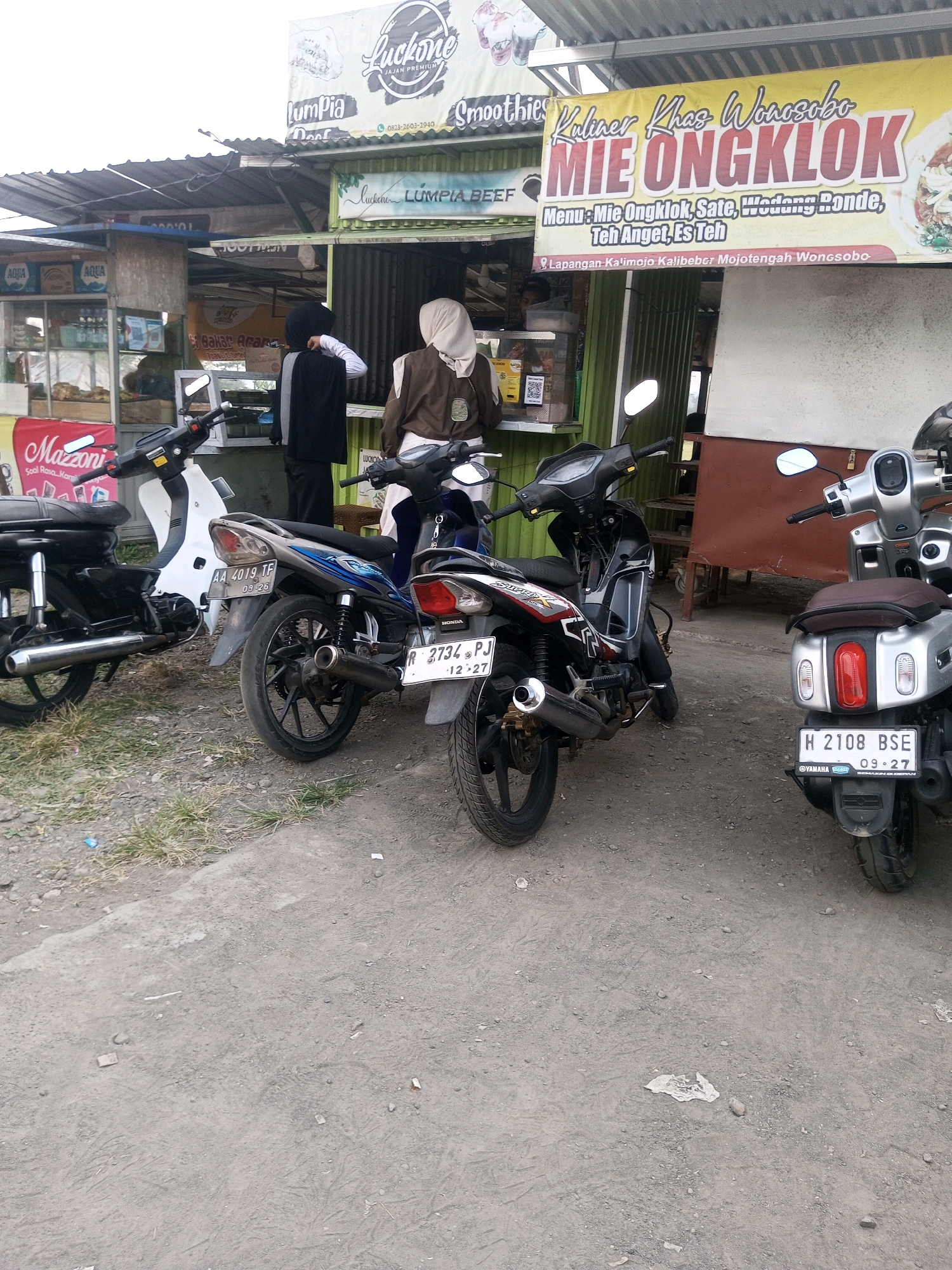 In Mojotengah, Indonesia, a street scene unfolds with three motorcycles parked on a gravel and dirt surface. The motorcycles are positioned in front of an open-air food stall. One motorcycle, a black and white model, is partially visible on the left. To its right, a blue and black motorcycle is parked, facing left. On the far right, a silver scooter is parked, facing right.
Behind the motorcycles, the food stall displays various signage. A prominent yellow banner advertises "MIE ONGKOK" and lists menu items such as "Mie Ongklok, Sate, Wedang Ronde, Teh Anget, Es Teh." The banner also indicates the location as "Lapangan Kalimojo Kalibeber Mojotengah Wonosobo." Two individuals are visible at the stall. One, wearing a black hijab and a gray and black jacket, is interacting with a person inside the stall. Another person, wearing a light brown hijab and a brown jacket, stands behind the blue and black motorcycle, looking towards the stall. Behind them, a white structure suggests a building or awning. The environment appears to be an outdoor setting, possibly a roadside market or food court, with a slightly overcast sky.