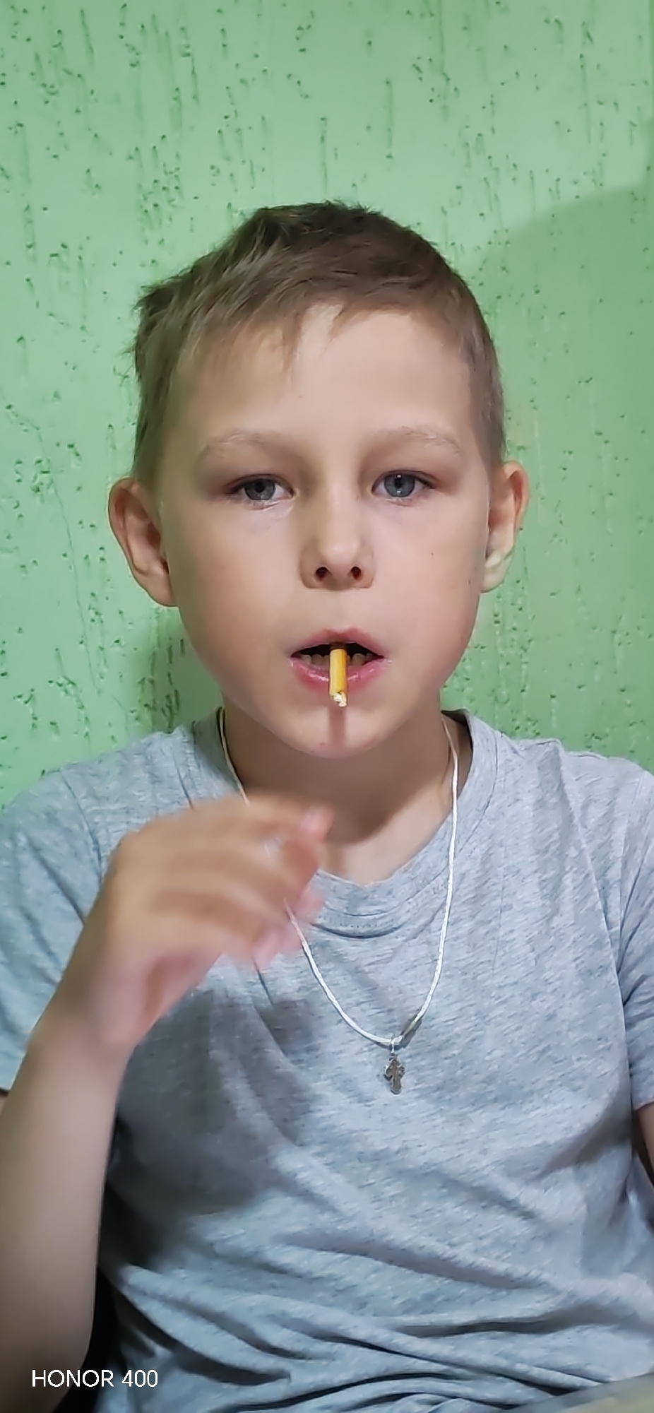A young fair-skinned boy with light brown hair and blue eyes looks directly forward. He is wearing a light grey short-sleeved t-shirt and a thin silver-colored chain with a small crucifix pendant around his neck. A small, light-brown cylindrical snack stick is held vertically between his upper and lower front teeth. His left hand is partially visible on the left side of the frame, slightly raised. The background is a uniformly light green wall with an irregular, textured surface. The device watermark "HONOR 400" is present in the bottom left corner of the image.