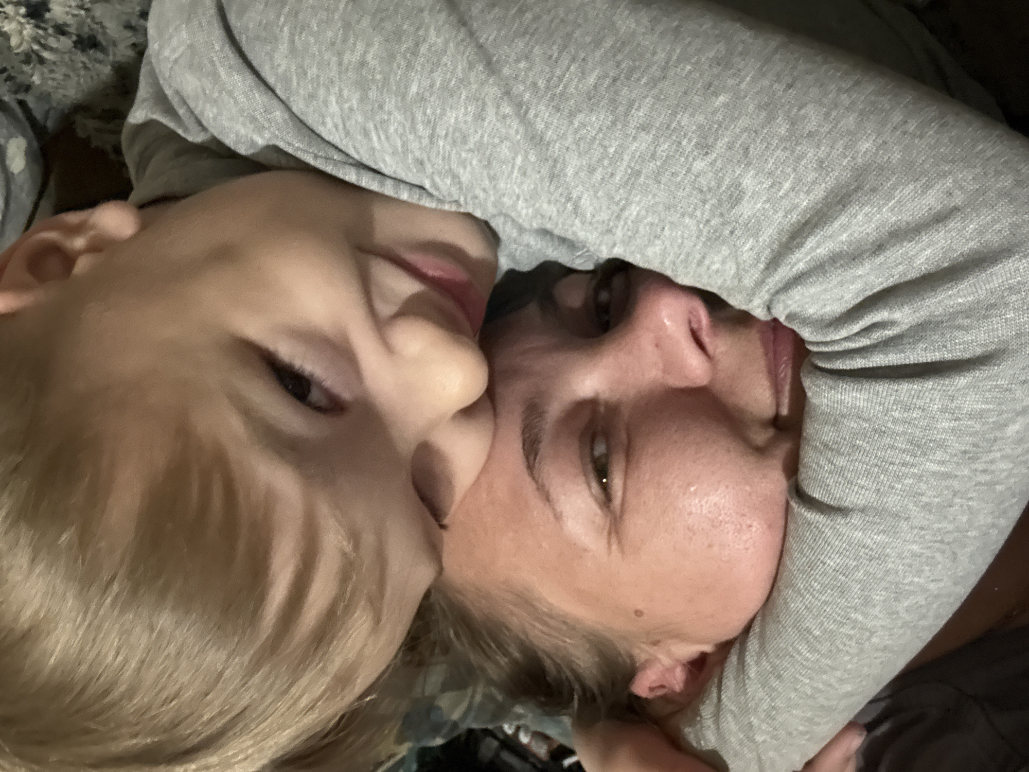 A photo captures a close-up view of a child and adult hugging, presumably lying down. The child, positioned on the left, has blond hair and is leaning into the adult, who is on the right. The adult has lighter hair, possibly also blond or light brown. Both are facing the camera, though the angle and proximity of the subjects make it difficult to discern their expressions. A grey long-sleeved garment is draped around the adult, possibly belonging to the adult or child. The background is indistinct, but what is visible looks like clothing or bedding. The location is not determinable to be in Dmitrov, Russia from the image.
