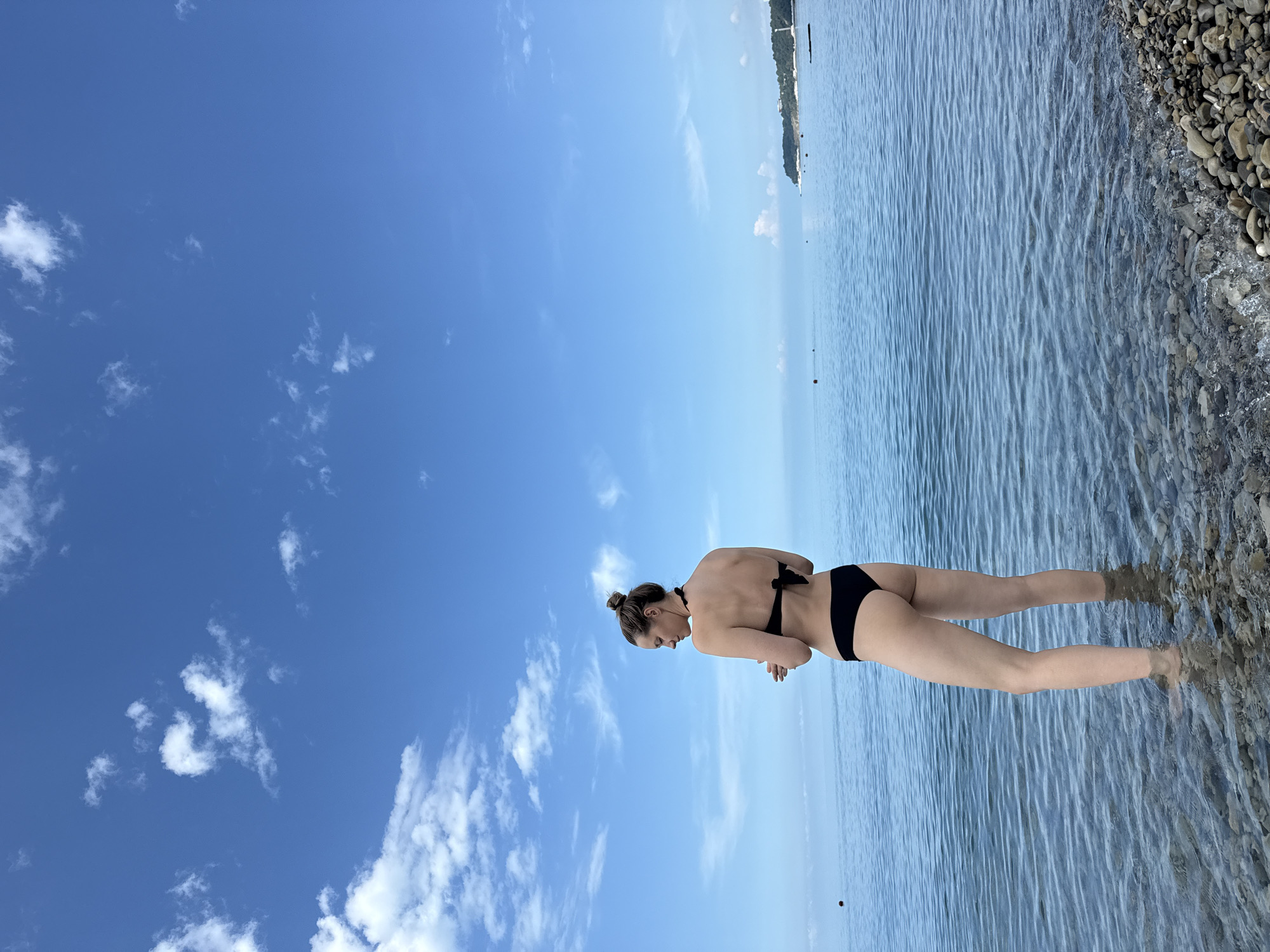 A woman stands in shallow, clear water on a pebble beach, facing away from the camera. She is wearing a black bikini. The sea extends to the horizon under a bright blue sky with scattered white clouds. In the distance, a coastline with trees and buildings is visible. The scene appears to be the Black Sea coast in Nebug, Russia, given the natural elements and the type of shoreline.