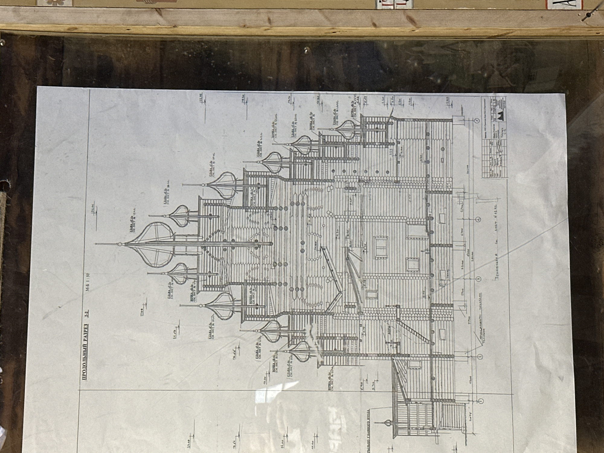 The image displays a detailed architectural drawing, likely a technical blueprint, of a wooden building with multiple onion domes, characteristic of Russian church architecture. The drawing is a longitudinal section, indicated by the Russian text "ПРОДОЛЬНЫЙ РАЗРЕЗ" at the top left, and a scale of "М-Б 1:50". The blueprint is mounted on a board behind glass, with some glare visible. The lower right corner of the blueprint contains a title block and other technical notes, including reference to "г. Невский Парклесхоз, Россия" (Nevskii Parkleskhoz, Russia), suggesting the location of the project. The drawing itself is in black and white and shows intricate details of the building's construction, including logs, windows, roofs, and decorative elements. There are no people visible in the image. The environment appears to be an indoor exhibition or archive, given the framing and lighting.
