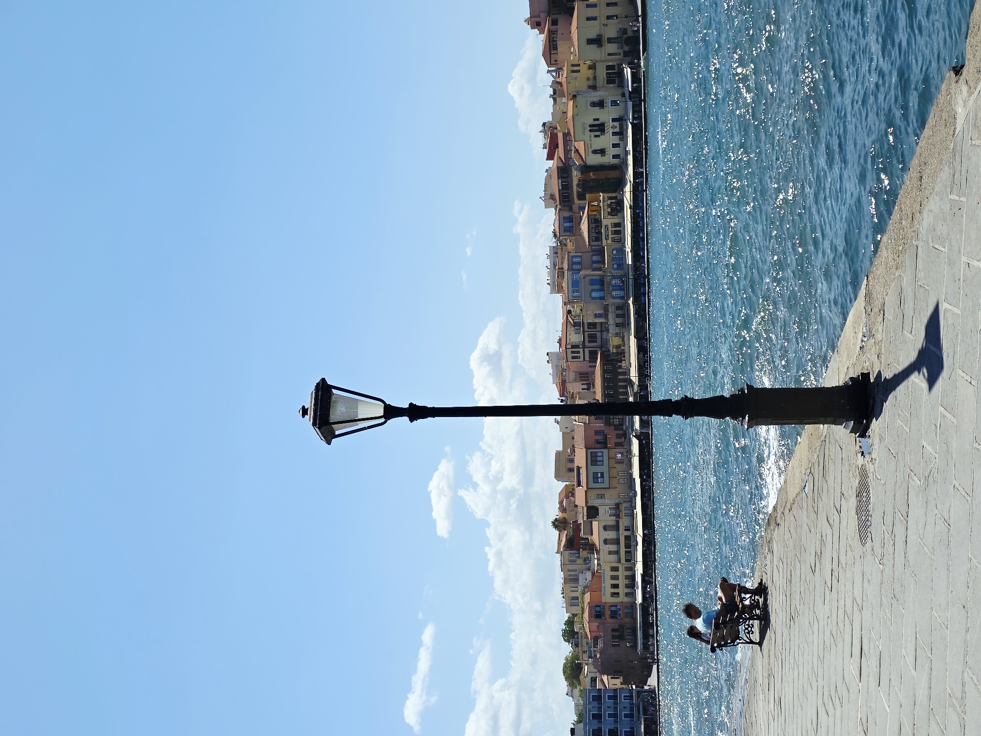 In Chania, Greece, a paved walkway runs along the waterfront with the sea on the right and buildings in the distance on the left. A lamppost stands on the walkway, casting a shadow. Two people sit on a bench facing the sea, with one person reading. The water is a vibrant blue, with sunlight glinting off the surface. The buildings are a mix of colors and architectural styles, with awnings visible along the waterfront. The sky is a clear blue with a few white clouds.