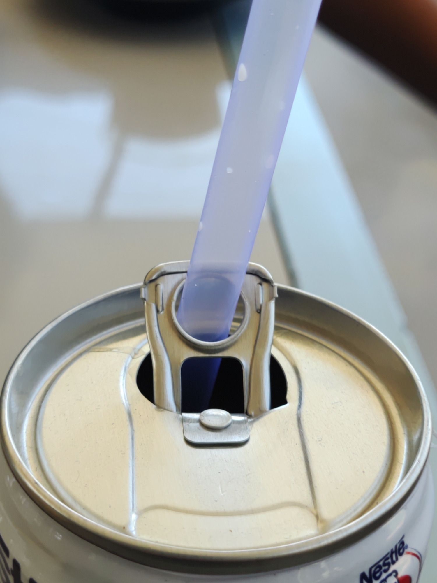 The image provides a close-up, high-angle view of a beverage can with a translucent purple straw inserted into its opening. The can, likely made of aluminum, features a standard pull-tab lid, which is pushed back to hold the straw firmly in place. The straw itself has several small, faint white specks on its surface.
The can is predominantly silver on top and white on its visible side, where a portion of the "Nestle" logo is clearly discernible in dark blue lettering. The object rests on a light-colored, possibly reflective surface, which is largely out of focus. The background is blurred, showing soft transitions between lighter and darker areas, suggesting an indoor setting.
The activity depicted is the act of drinking or preparing to drink from a canned beverage using a straw. There are no people visible, so emotions cannot be inferred. The lighting appears bright and even, not indicating a specific time of day. Given the provided context, the scene is located in Lhokseumawe, Indonesia.