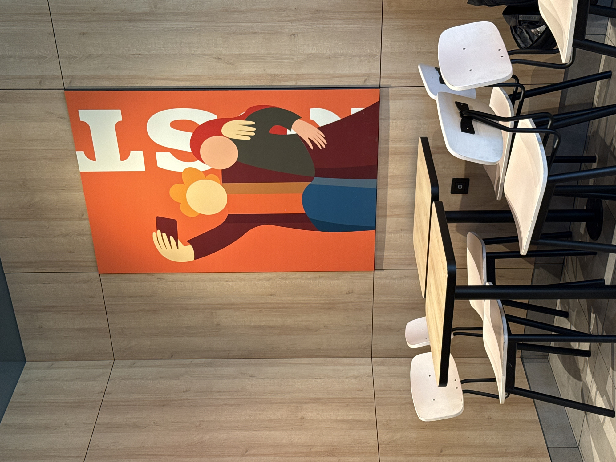 The image depicts a corner of a modern, indoor establishment, likely a cafe or restaurant. The walls are paneled with light-colored wood laminate, creating a warm, natural ambiance. A large, vibrant orange art piece adorns one wall, featuring a stylized depiction of two figures holding each other and taking a selfie with a smartphone. The figures are rendered in bold, flat colors, with one person having a yellow, flower-like head and the other wearing a red headscarf.
In the foreground, several minimalist tables and chairs are arranged. The tables have light wood tops and black metal legs, with a modern, slightly rounded square design. The chairs are also minimalist, with light-colored wooden seats and backs, and thin black metal legs. Some of the chairs are empty, while others appear to be occupied by a person or persons not fully visible. The floor is tiled in a neutral gray tone. The overall impression is one of a clean, contemporary space designed for casual dining or socializing. The establishment is located in Izhevsk, Russia.