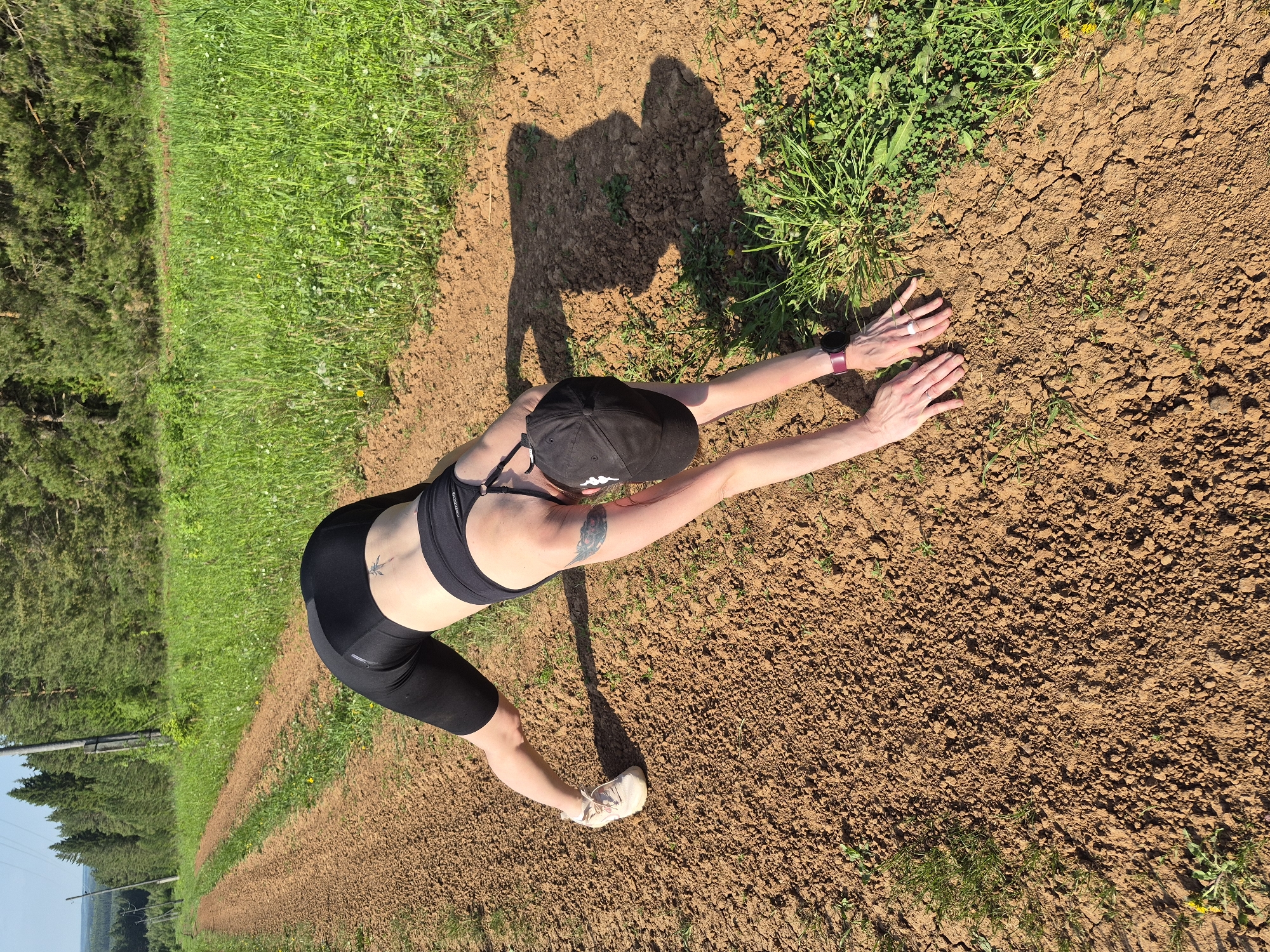 A female wearing a black sports bra and shorts, a black baseball cap, and white sneakers is stretching on a patch of brown dirt adjacent to a grassy area. The scene is bathed in sunlight, casting a shadow of the woman on the ground. The location appears to be rural, with trees visible in the background.