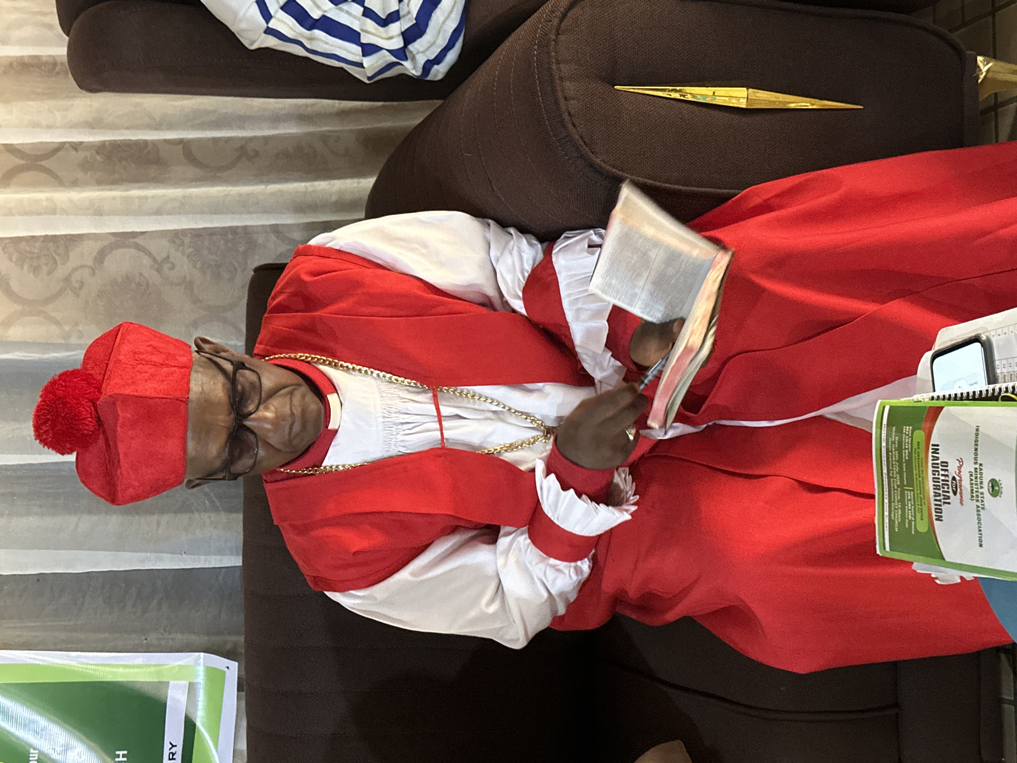 A Black man wearing a red and white clerical robe and a red clerical hat with a pom-pom sits on a brown couch, holding an open book. He is wearing glasses and a gold chain. To his right, another person in a blue and white striped shirt is partially visible. Behind the couch is a light-colored curtain with a floral pattern, and to the left of the man is a green banner with white text. A smaller green booklet lies on the couch in front of the man, and a yellow decorative object is visible on the back of the couch. The scene appears to be indoors, possibly in a church or community hall, given the presence of the banner and booklet. This event is located in the city of Barden Yana, Nigeria.