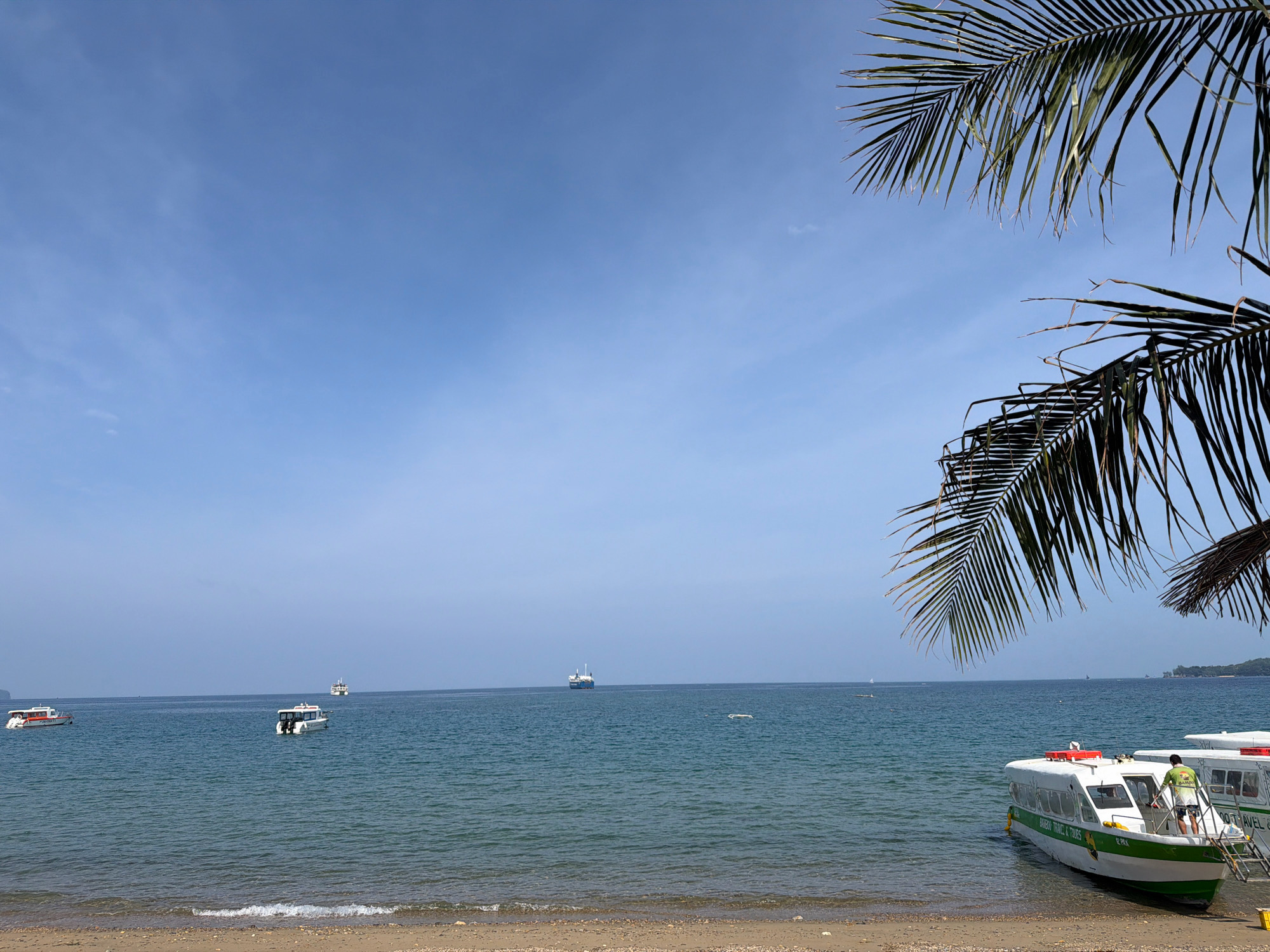 The image depicts a clear, sunny day at a beach in Malay, Philippines. The foreground features a sandy shore with small pebbles and a gentle wave washing onto the beach. Two white speedboats with green accents are partially beached on the sand, facing the water. A person wearing a green t-shirt and shorts is standing on the ladder of the boat closer to the viewer, appearing to be preparing for departure or disembarking. Another boat is visible behind it, partially obscured. In the calm, blue sea, several other boats are scattered in the distance, including smaller speedboats and what appears to be a larger ferry or cargo ship further out. Palm fronds from trees overhang the scene from the top right, suggesting a tropical location. The sky is a clear, pale blue with minimal cloud cover.