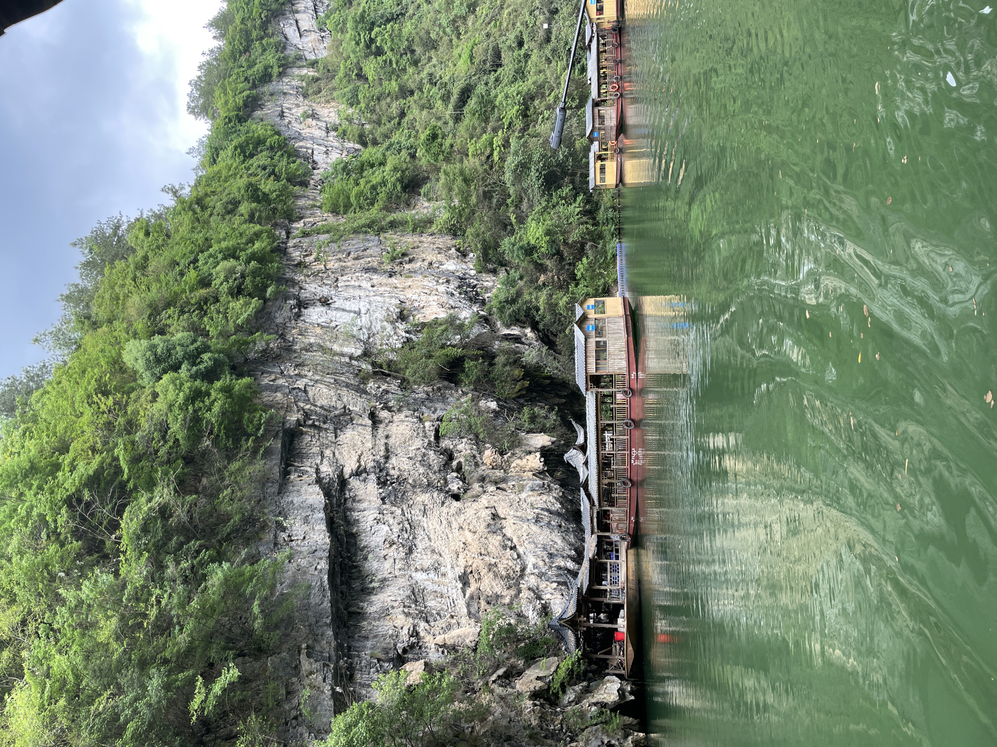 The scene depicts a riverbank in Sanguansi, China. A sheer rock face rises vertically, covered with vegetation, including trees and shrubs. Moored along the water's edge are several multi-story boathouses. The water reflects the structures and the surrounding landscape. The water is colored green.