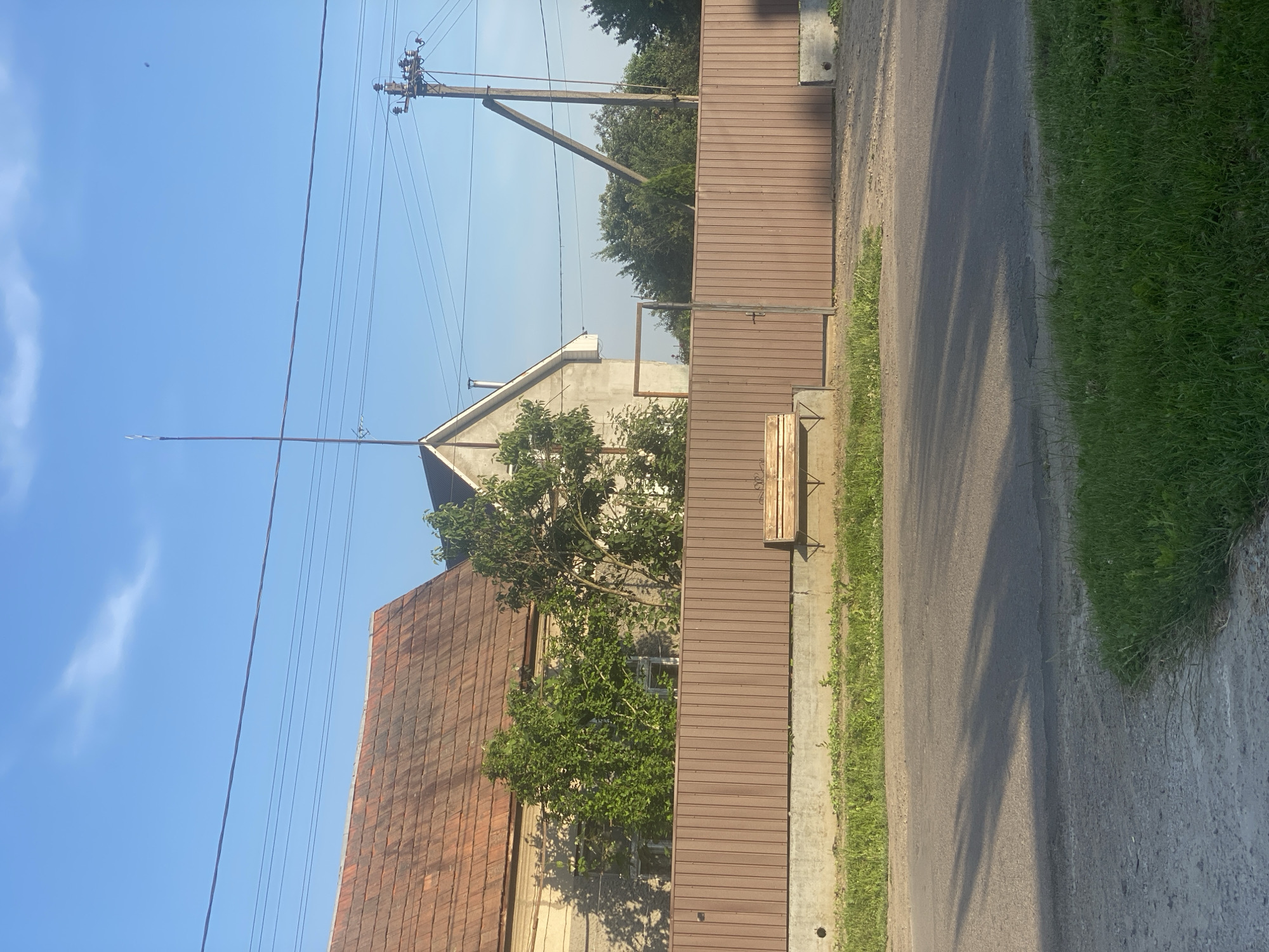 A view of a street in an undetermined location shows a brown metal fence that stretches across the frame. To the left of the fence, a portion of a building with a reddish-brown tiled roof and beige walls is visible. Two trees with green foliage stand behind the fence, casting shadows on the building. In front of the fence, a simple wooden bench rests on a concrete foundation. A small gate is integrated into the fence, with a thin vertical metal bar. The road in front of the fence is asphalt, with tire tracks and shadows cast by unseen objects. Grass and weeds grow along the edge of the road and in front of the fence. Overhead, power lines and a utility pole are visible against a clear blue sky with a few wispy clouds.
