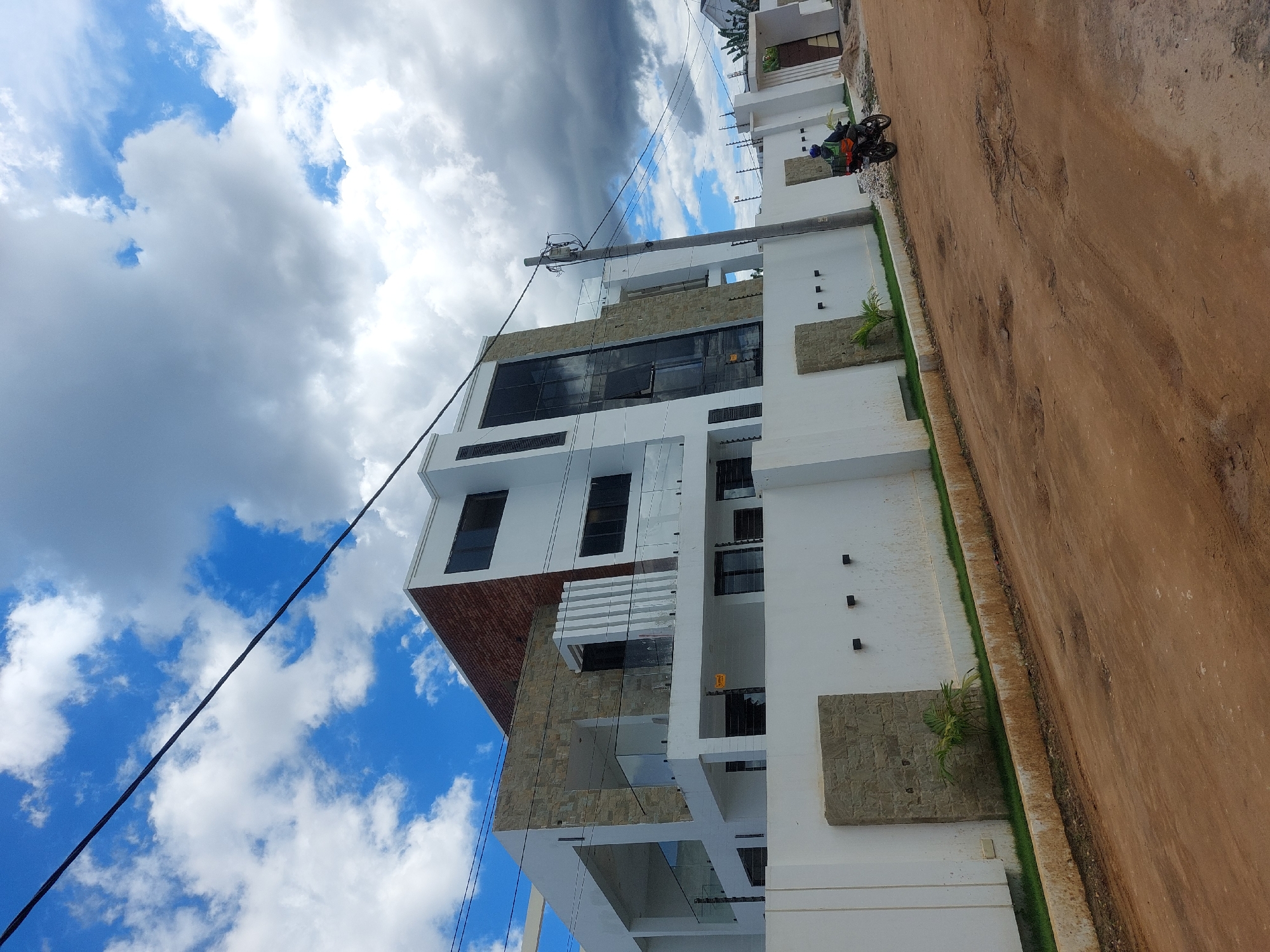 The image depicts a modern, multi-story building with a white facade and stone accents, potentially a residential or commercial structure. Dark-tinted windows are present. The building is located alongside a dirt road, with a patch of grass separating the building from the road. A motorcycle with two riders is visible in the vicinity of the building. Power lines are seen running near the building. The sky is filled with a mix of blue sky and white, billowing clouds, suggesting either a sunny or partly cloudy day.
Given the architecture and environment, it can be suggested that this building is likely located in a developing area within Dar es-Salaam, Tanzania.