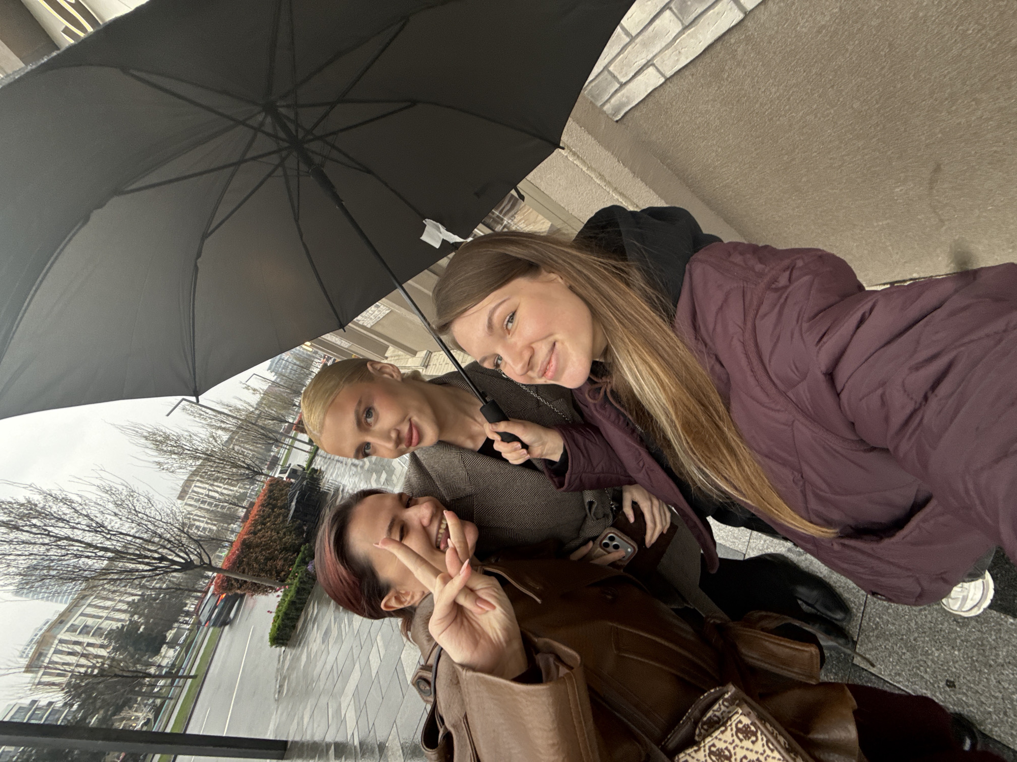Three young women are posing for a selfie under a black umbrella on a wet city street. The woman in the foreground has short red hair and is wearing a brown leather jacket, making a peace sign. The woman to her right is blonde and wearing a brown jacket and holding a cell phone. The third woman, behind the blonde, has long brown hair and is wearing a plum-colored jacket. Buildings, trees, and a street with a car are visible in the background. The photo was taken in Belgrade, Serbia.