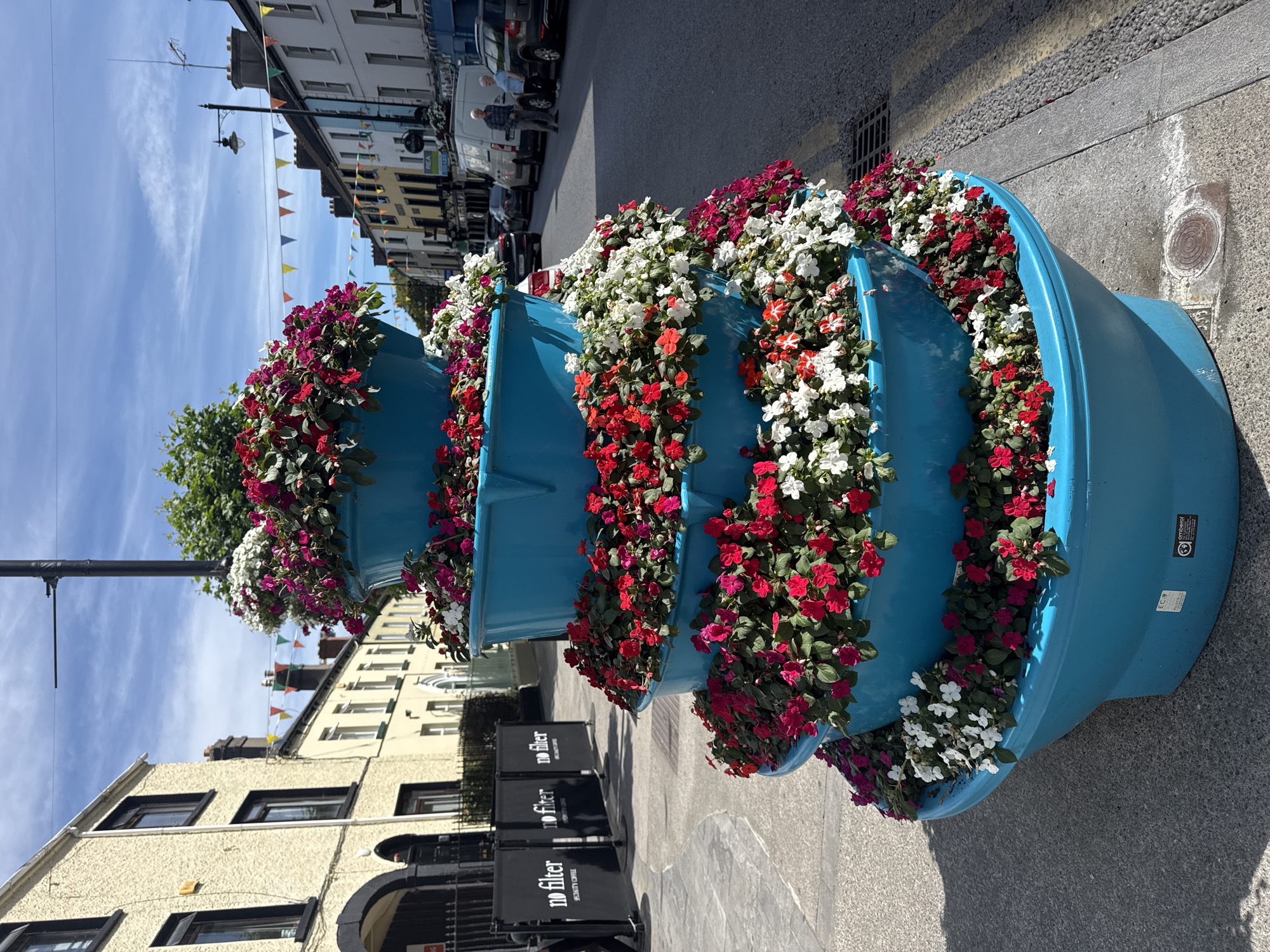 This is a medium-shot, eye-level, outdoor, daytime photograph of a large, tiered, blue planter filled with an abundance of red, white, and pink impatiens flowers. The planter is situated on a paved sidewalk next to a street in Clonmel, Ireland. The flowers cascade over the edges of each tier, creating a voluminous display.
In the background, to the left, is a building with a pale yellow facade, dark trim around windows and doorways, and a black metal fence. Further down the street, on the right, are several two-story buildings with colorful facades, including light blue, yellow, and white. A row of triangular flags in various colors (red, yellow, blue, green) is strung across the street between buildings. Several cars are parked along the street, and a white van is visible. Two people, an older man and an older woman, are standing on the sidewalk to the right of the van. A lamppost with multiple arms is visible above the planter. The sky is blue with scattered white clouds. The ground in the foreground is paved with grey concrete and cobblestones, and a metal drain cover is visible near the base of the planter. Labels on the bottom of the planter indicate it is from "amberol" and "all-eco."
