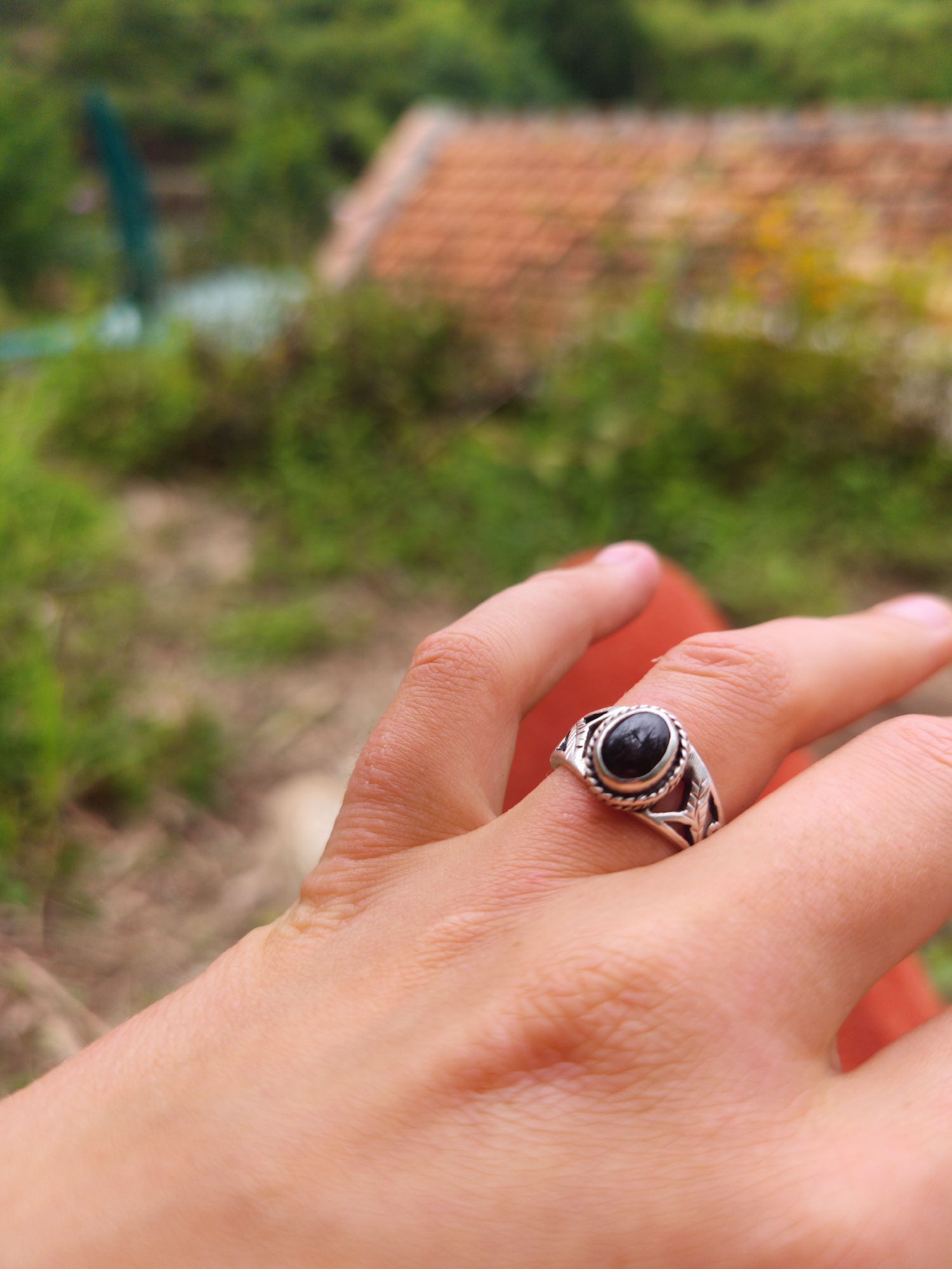 A photograph taken in what appears to be an outdoor setting, likely Cam Ly - Da Lat, Vietnam, focuses on a human hand wearing a silver ring with a black stone. The ring is on the middle finger. The background is blurred, but shows greenery and a building with a reddish-brown roof.