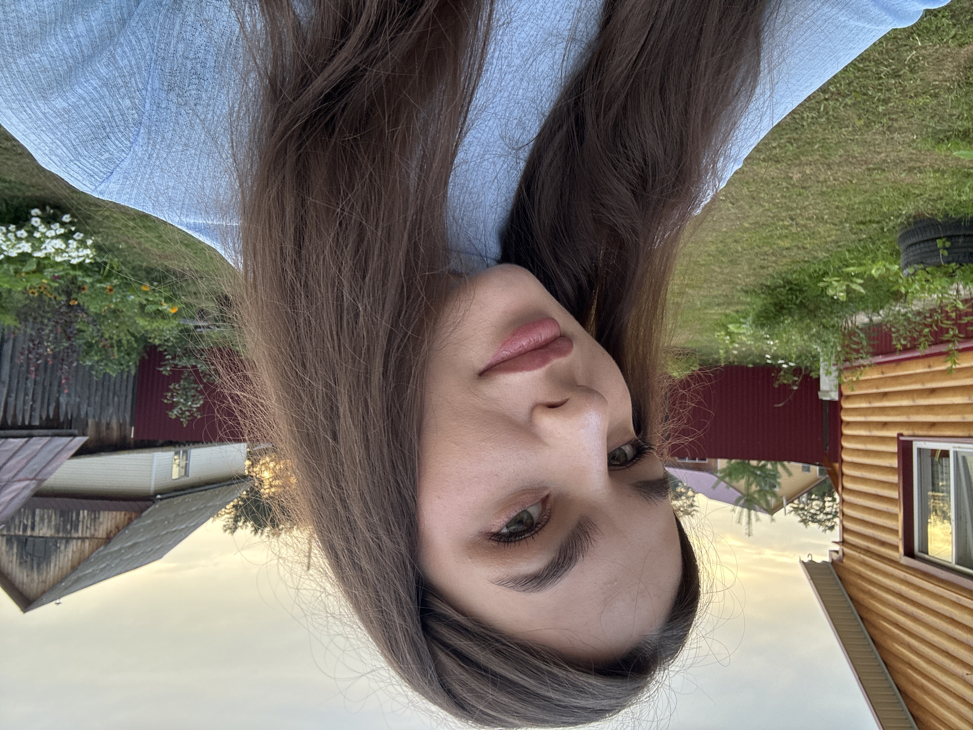A young woman with long, brown hair and fair skin takes a selfie outdoors. She is wearing a light blue, textured shirt and has on makeup, including eyeshadow and lip gloss. Her hair is loose and frames her face. The background shows a rural setting with a wooden house on the left, a red fence, and other houses and vegetation in the distance. The sky is overcast with hints of sunset light. The scene appears to be in Rusalevka, Russia, likely a village or dacha settlement, given the typical architecture and landscaping.